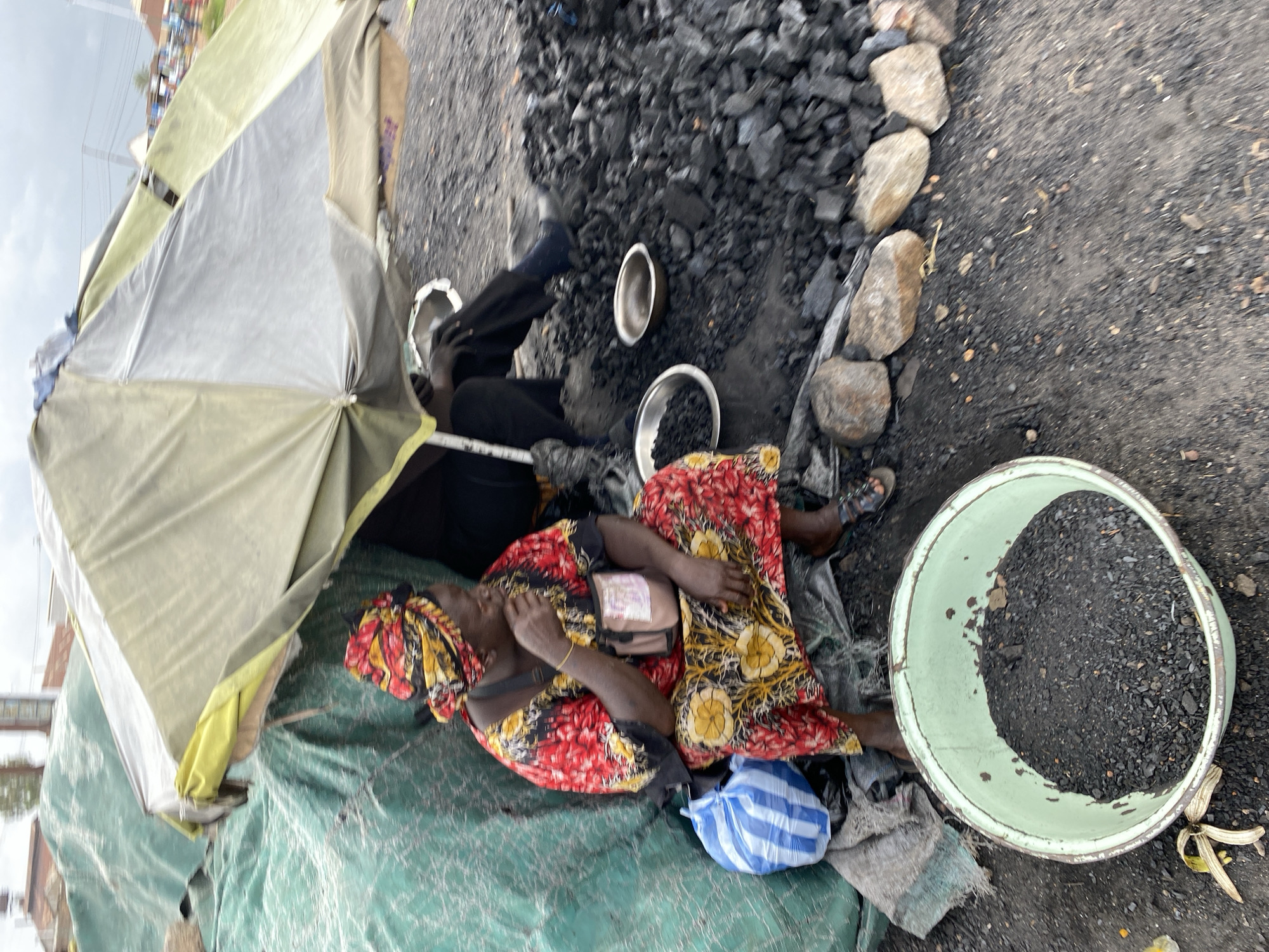 In Bukuru, Nigeria, a woman sits under a makeshift shelter, a vibrant floral dress and matching headwrap adorning her. She is surrounded by piles of charcoal, a staple for cooking and fuel. A large blue and white striped bag rests by her side, possibly containing supplies or goods for sale. Two shallow metal bowls, one filled with charcoal, sit on the ground before her, suggesting she is either selling the fuel or preparing it for use. The scene unfolds outdoors, with a dusty, uneven ground littered with scattered charcoal and a few rocks. The shelter, constructed from tarps and an umbrella, offers protection from the elements, indicating a market or roadside stall setting. The overall impression is one of everyday commerce and survival in a bustling Nigerian city.