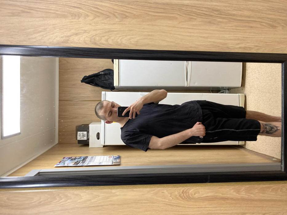 A male individual is depicted in a mirror selfie taken indoors in Марфино, Russia. The man, with short buzzed hair and light facial stubble, wears a black short-sleeved t-shirt and black athletic shorts with a grey stripe on the side. A dark-colored tattoo, resembling a stylized skull or monstrous face, is visible on his left lower shin. He holds a black-cased smartphone in his right hand, angled downwards, capturing his reflection.
The environment reflected in the mirror features light-colored wood paneling on the walls. A rectangular white LED light fixture is mounted on the ceiling. On the left wall, a multi-page calendar is affixed. Two white upright refrigerators are present; a brown cardboard box rests on the top of the left refrigerator, and a black drawstring backpack is on top of the right refrigerator. The floor appears to be a light beige or tan material. The mirror itself has a dark, thin frame and occupies the majority of the foreground.