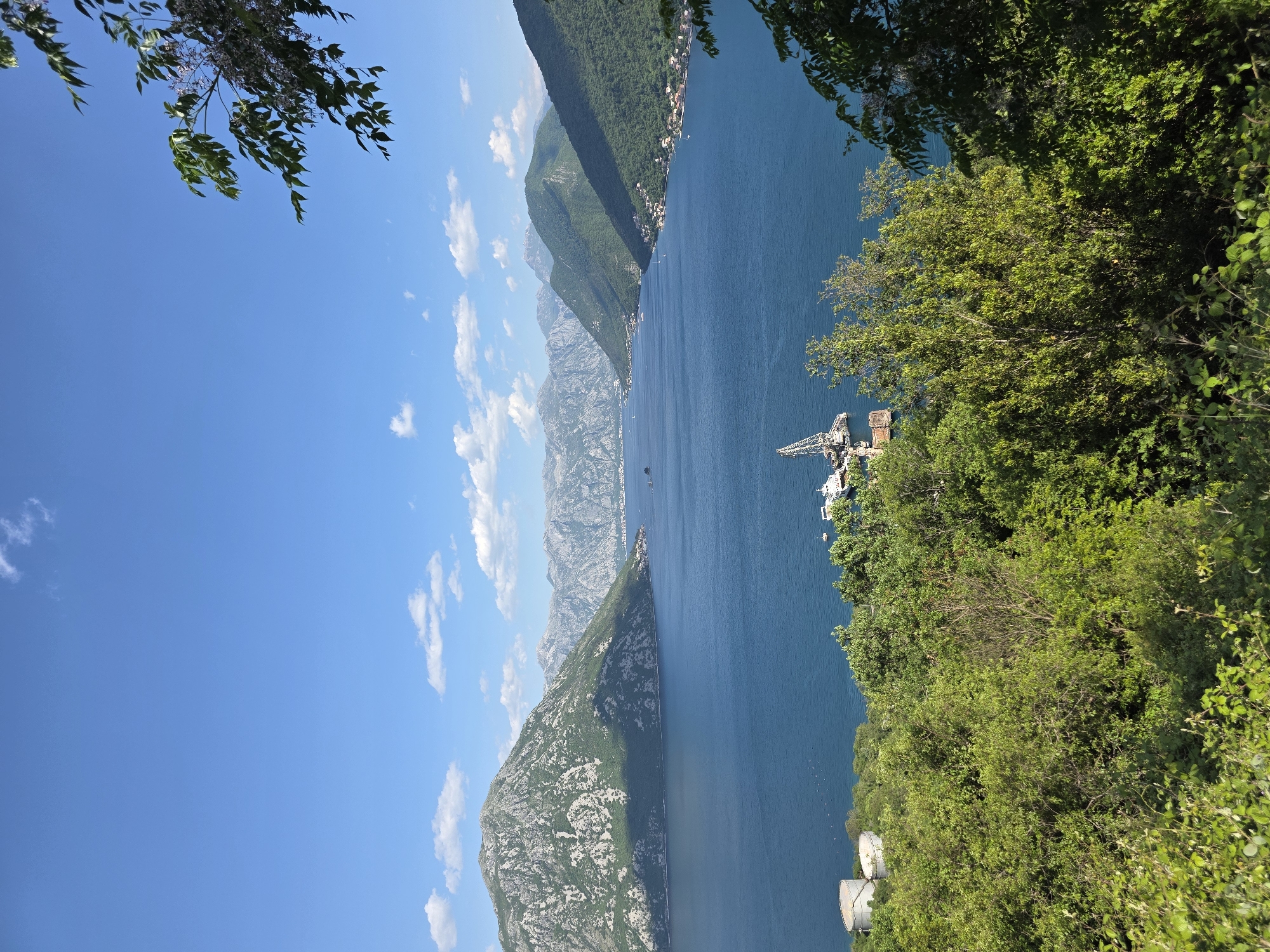 The image depicts a scenic view in Morinj, Montenegro. It is a bay surrounded by mountains. The water appears deep blue and contrasts with the light blue sky. Fluffy white clouds are scattered across the sky. There are several boats visible on the water, and a crane structure is positioned on the shore. Lush green foliage is seen in the foreground, framing the view.