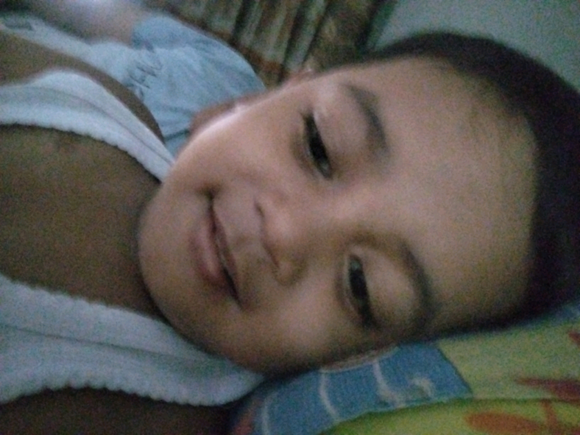 A close-up, overhead shot shows a sleeping child on their side. The child's face is turned towards the viewer. The child is lying on a blue and green patterned blanket, with a white blanket or pillowcase visible on the left side of the frame. The background consists of a brown, patterned surface, possibly a wall or curtain. The lighting is dim. The image was taken in Pepelegi, Indonesia.