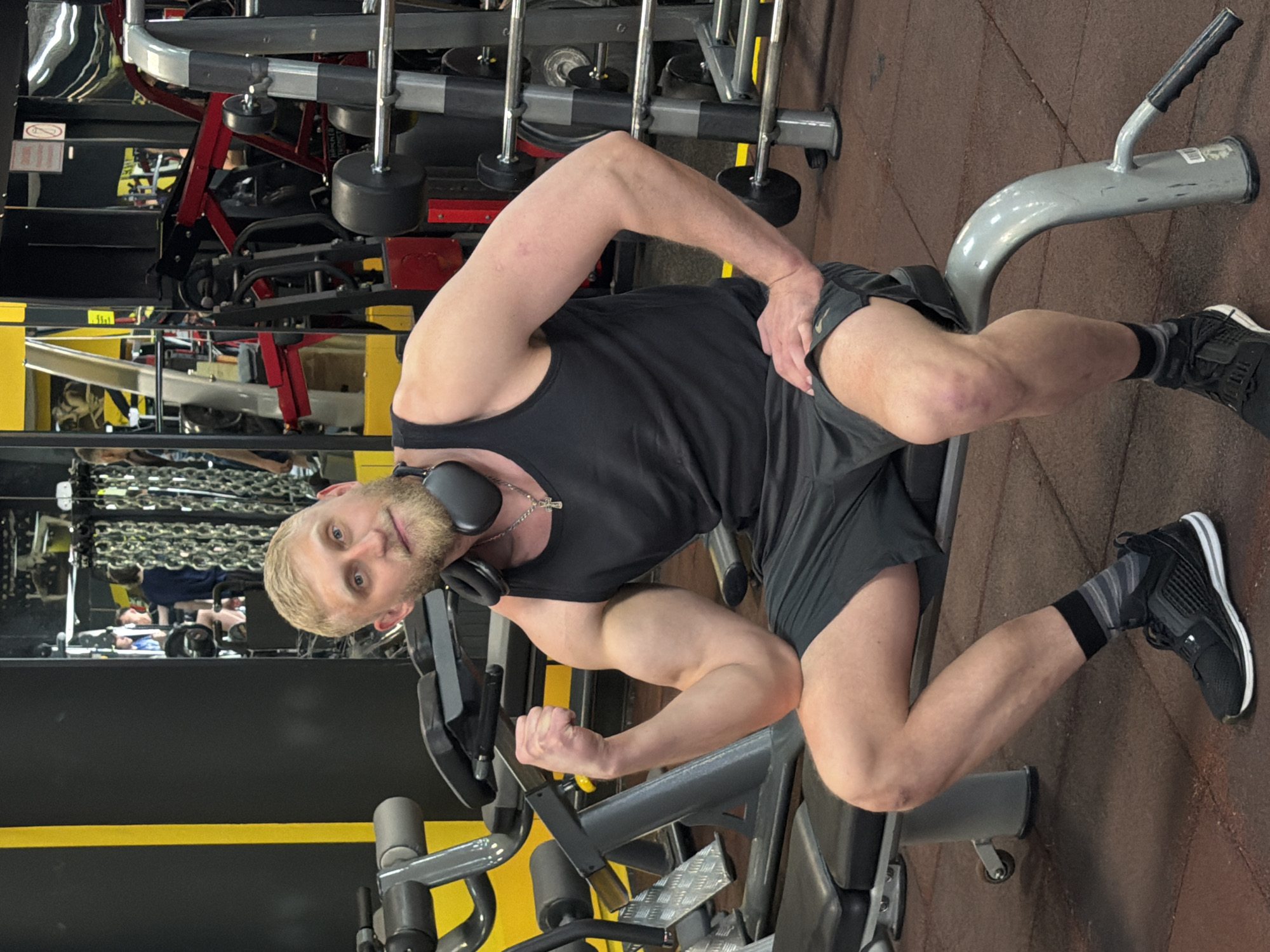 The image shows a muscular man with a blonde beard and short hair, wearing a black tank top and gray shorts, posing with flexed biceps while sitting on a leg extension machine in a gym. He has headphones around his neck and a silver chain with a cross pendant. He wears black sneakers with gray and black socks. The gym environment includes various weight machines with yellow and gray accents. The floor is brown and appears to be made of rubber. There is a mirror behind the man, reflecting gym equipment and other people exercising. The location is most likely in a gym in the city of Krasnoyarsk, Russia, but it is unconfirmed.
