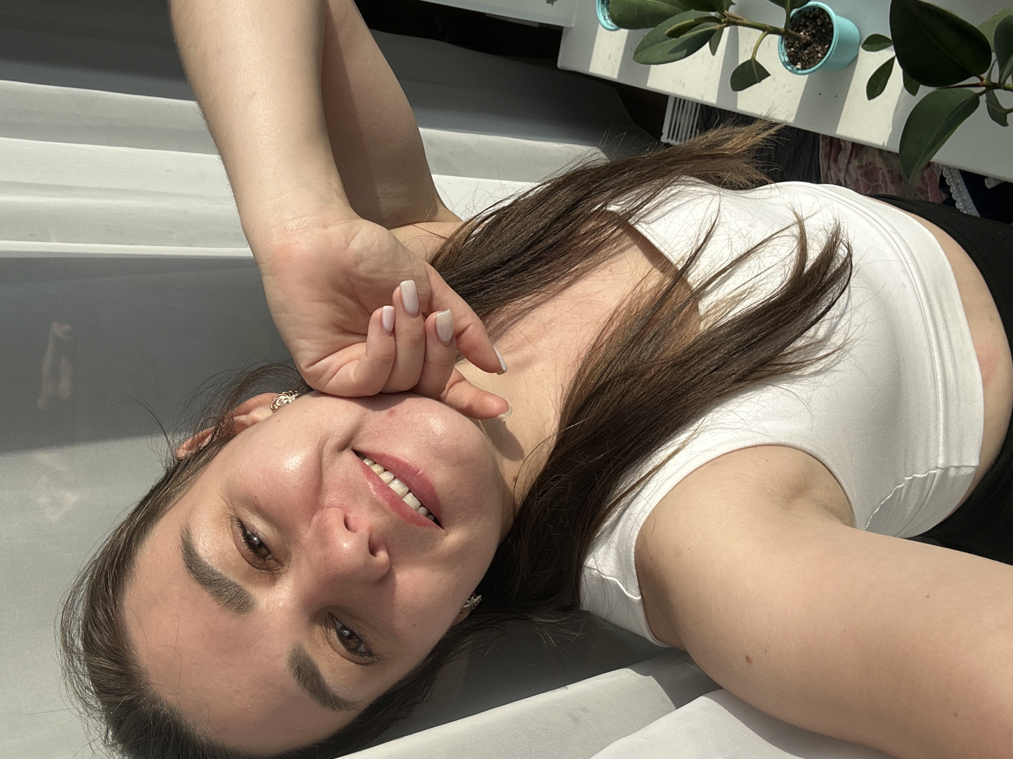The image shows a young woman lying on her back, looking up at the camera. She has long, dark brown hair and is wearing a white crop top. Her left arm is bent at the elbow, with her hand resting near her face. She is smiling and her eyes are open. The scene is outdoors, with a bright, sunny day suggested by the strong shadows and highlights. In the background, there is a white surface, possibly a railing or ledge, and a potted plant with green leaves. The overall impression is one of a casual, relaxed moment. The location is identified as Chaikovsky, Russia. There is no visible text in the image.