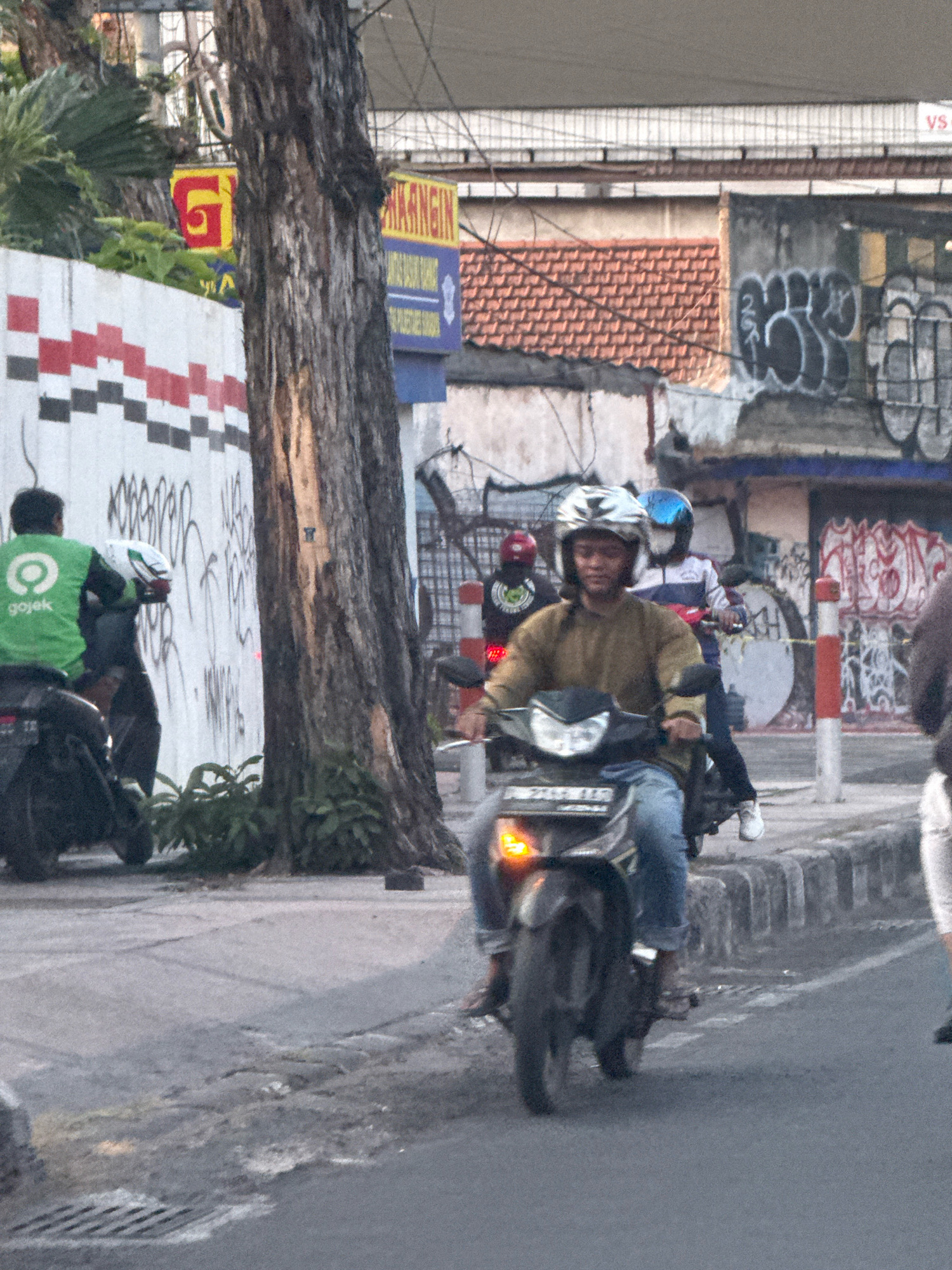 The image depicts a street scene in Surabaya, Indonesia, likely in a commercial or residential area. In the foreground, a person wearing a brown jacket and blue jeans is riding a motorcycle, looking towards the camera. They are wearing a helmet with a white and silver design. Behind them, another person on a motorcycle wears a blue helmet and a white and blue jacket. To the left, a person in a green Gojek jacket is also on a motorcycle, facing away from the camera. The motorcycles are on a paved road next to a sidewalk. Behind the sidewalk, there's a wall with a red, white, and black patterned design and graffiti. A large tree trunk is prominent in the center of the image, obscuring parts of the background. Further in the background, there are buildings with tiled roofs and more graffiti. Several signs are visible, including a yellow sign with a red "G" and a blue sign with text that is partially obscured. The overall lighting suggests it is daytime.