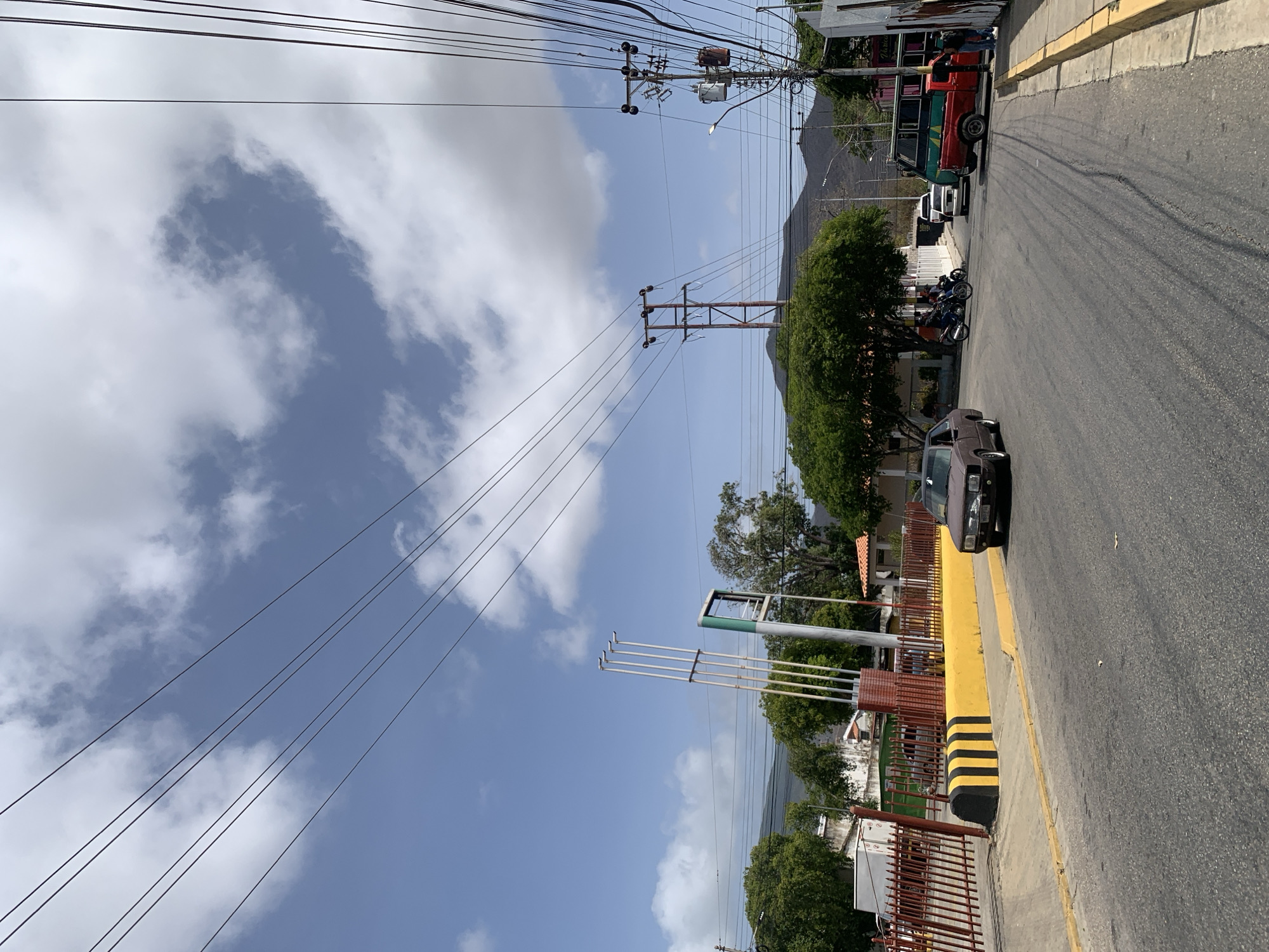 A street view in Santa Ana, Venezuela, presents a road bordered by a yellow curb. A black car is parked on the street. Power lines crisscross above, converging near utility poles. In the background, a mountain is visible. Several motorcycles and a green and red bus are parked further down the road. The sky is partly cloudy. A sign with multiple poles is present on the side of the road. A red metal fence borders the sidewalk.