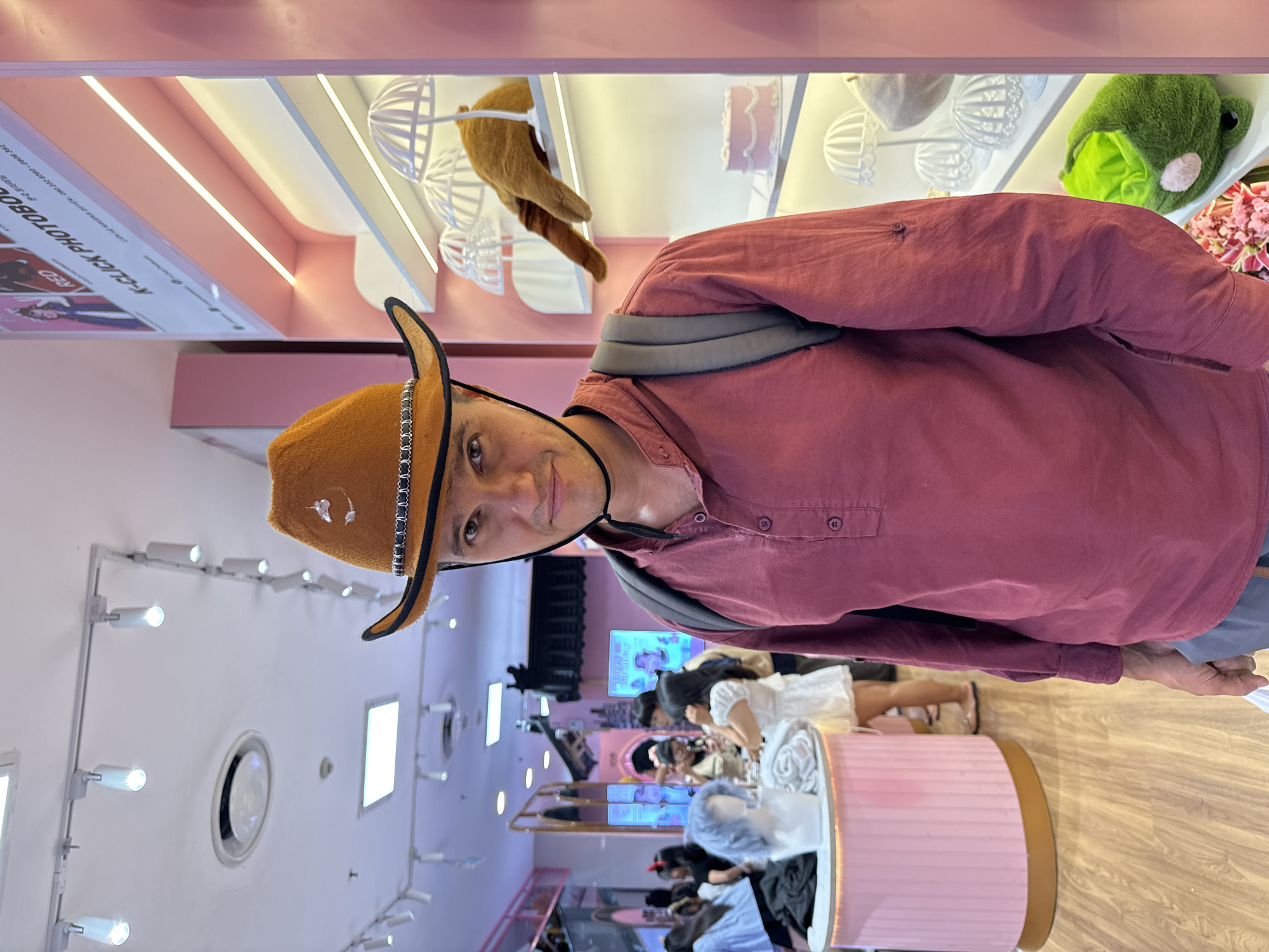 A medium shot, slightly high-angle perspective, features a man wearing a brown cowboy hat and a maroon long-sleeved shirt, inside what appears to be a brightly lit shop or attraction, possibly in Phú Thủy, Vietnam. The man, with a backpack strap visible, is looking at the camera. Behind him, the background is a store with a pink aesthetic, showcasing various headwear including hats shaped like animals and other novelty designs on shelves. Other customers are visible in the background, some seated. The foreground includes a pink and tan display. The environment is brightly illuminated with strip lights. The image composition is slightly off-center.