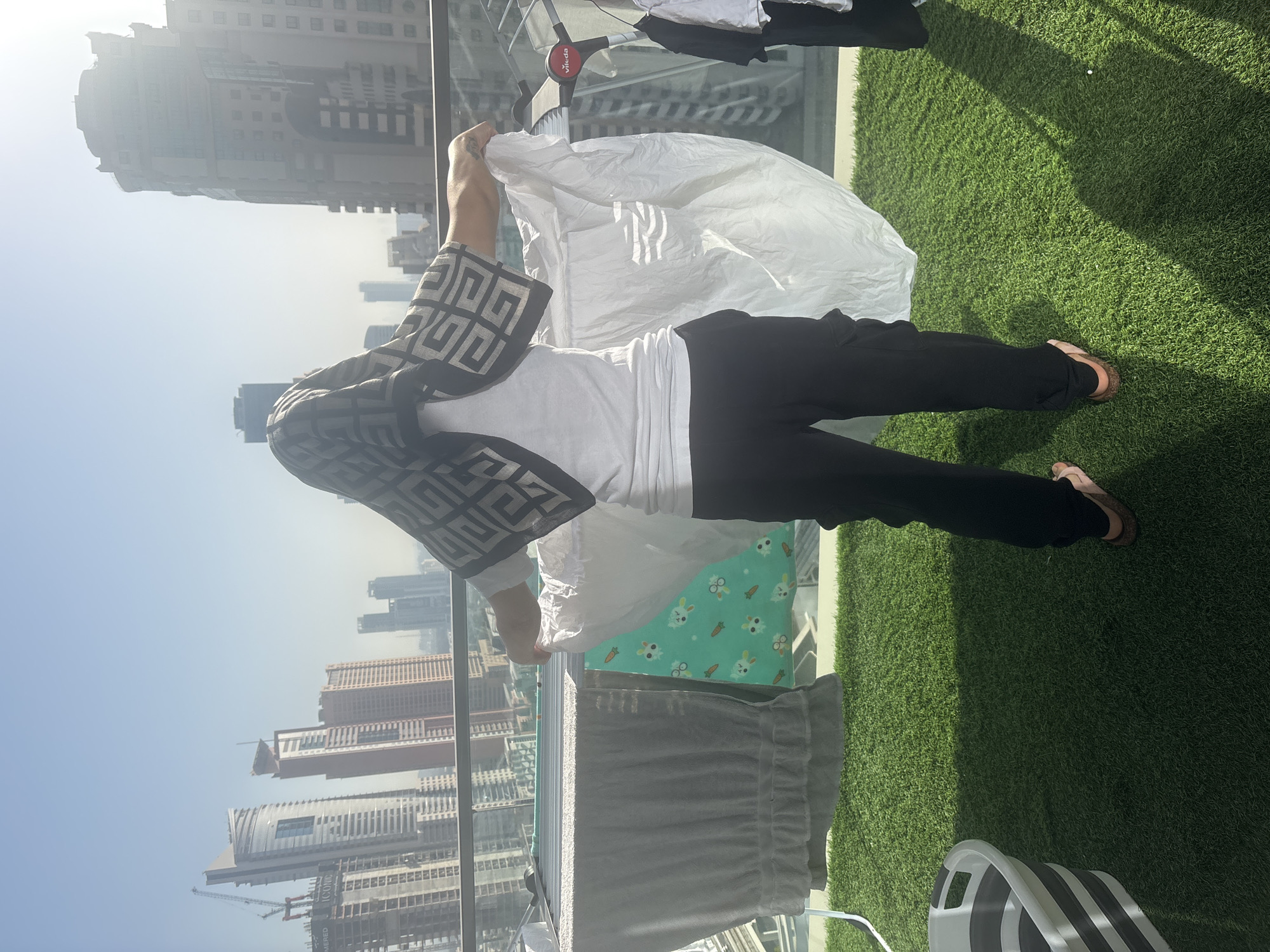 A person stands on a balcony covered in artificial turf, with a sprawling cityscape of Dubai visible in the background. The person is wearing black pants, a white t-shirt, and a patterned headscarf. They are hanging laundry on a drying rack, holding a large white sheet. Adjacent to the drying rack, a grey and white striped laundry basket sits on the artificial turf. The sky is clear and bright, suggesting a sunny day. The buildings in the background are a mix of modern high-rises, some under construction.