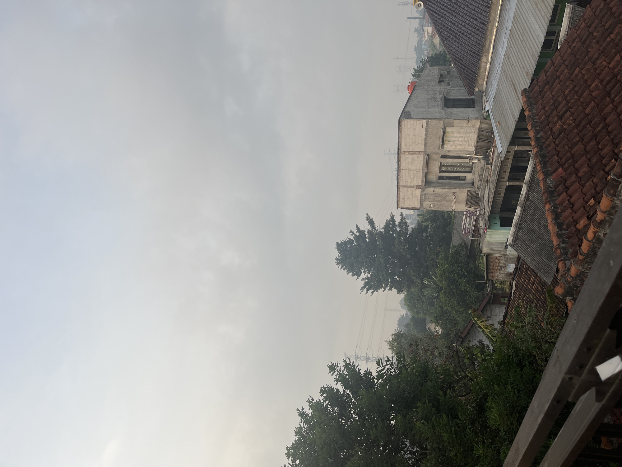 The image captures an overcast sky over a densely populated urban area. In the foreground, a metal railing overlooks a cluster of rooftops, primarily made of terracotta tiles and corrugated metal. Beyond these, various buildings are visible, some appearing partially constructed with exposed brickwork, while others are complete with windows and doors. Trees with dense green foliage are interspersed throughout the scene, particularly on the left side and towards the middle, obscuring some of the structures. In the background, power lines stretch across the hazy sky, supported by tall transmission towers, indicating the presence of electrical infrastructure. The overall impression is one of a residential neighborhood under a muted, possibly polluted, sky. The location is identified as Pasir Gadung, Indonesia.