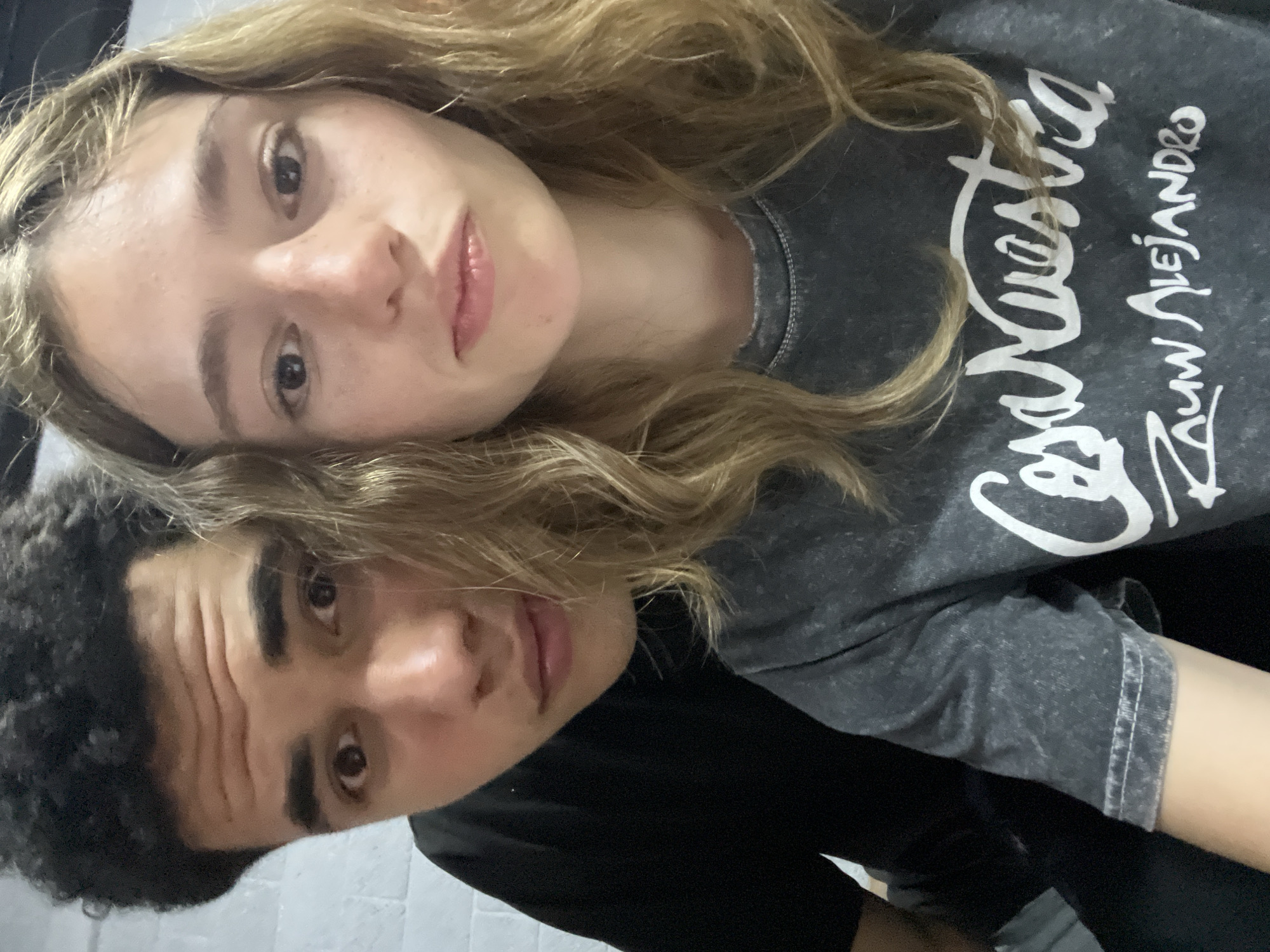 Two individuals, one female with blonde hair and one male with dark hair, are positioned closely together. The female is wearing a gray t-shirt with white lettering and the male is wearing a black shirt. The image appears to be taken indoors. The location is not indicated in the image and the presence of San Bernardino Tlaxcalancingo, Mexico cannot be confirmed.