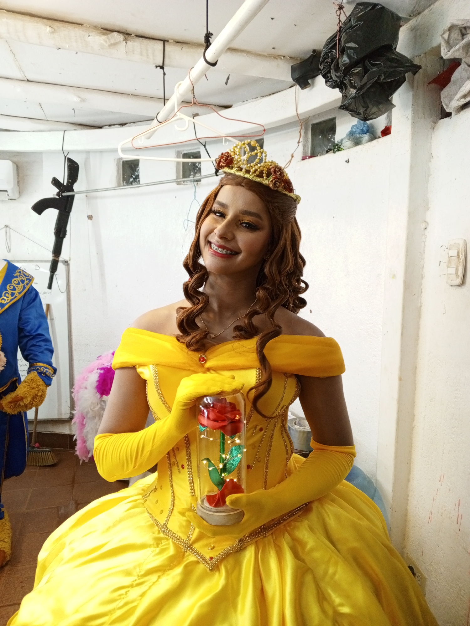 A young woman dressed as Belle from Beauty and the Beast stands smiling, holding a rose encased in glass. She wears a yellow gown with matching gloves and a gold crown. To her left, partially visible, is someone in a Beast costume. The location appears to be a cluttered backstage or storage area, with white walls and various items hanging from the ceiling or stored on shelves. This image was captured in Porlamar, Venezuela.