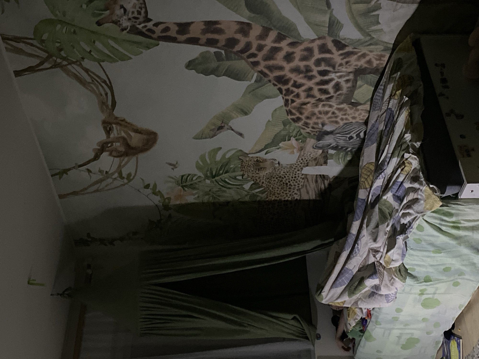 The image depicts a room with wallpaper featuring a jungle scene with animals, including a giraffe, leopard, zebra, and monkey. The room contains a bed with a green and white patterned bedspread and a draped green curtain. The location is Chaikovsky, Russia.