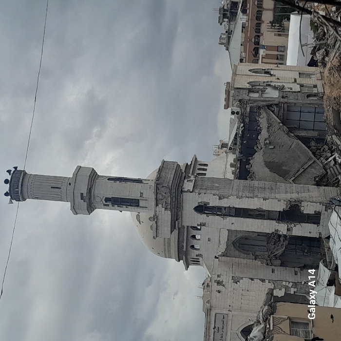 The image captures a severely damaged mosque in Khan Yunis, Palestinian Territory, under an overcast sky. The white minaret, though still standing, exhibits extensive structural compromise, featuring cracks, missing facade sections, and exposed interior elements across its octagonal base and cylindrical upper shaft. Several loudspeakers are mounted on the minaret's pinnacle. The mosque's main dome, situated behind the minaret, displays multiple circular impact marks. The primary building structure of the mosque is largely destroyed, with significant portions of its walls and roof having collapsed inward, revealing extensive rubble, twisted rebar, and dark interior spaces. Remaining sections of the white walls show numerous apertures where windows or other architectural features once stood. The immediate foreground and surrounding areas are covered with large piles of concrete, rebar, and other building debris. Utility wires are visible horizontally across the upper frame. No individuals are present in the scene. The watermark "Galaxy A14" appears in the lower left corner.