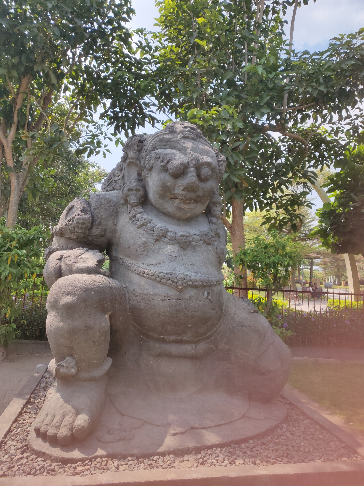 The image shows a large stone statue of a mythical creature, possibly a raksasa or a guardian deity, in an outdoor setting. The statue is grey and weathered, with detailed carvings of jewelry, hair, and facial features. It is seated with its left leg bent and its right leg extended. The statue is surrounded by lush green trees and foliage, with a blue sky visible through the leaves. In the background, a metal fence encloses a grassy area with a few people sitting on benches. The ground around the statue is covered in gravel and dirt. This statue is located in Pagu, Indonesia.