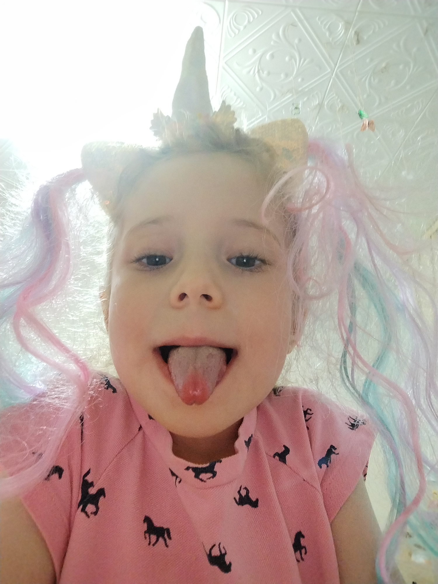 A young child, likely female, poses for a close-up, low-angle shot. She is wearing a unicorn headband with a gold horn and ears. Her hair is styled into pigtails with strands of pink and blue wig extensions. The child is wearing a pink shirt with a pattern of black horse silhouettes. She is sticking her tongue out. The background is a textured white ceiling. The location is unspecified, and there is no evidence to indicate this picture was taken in Марковский, Russia.