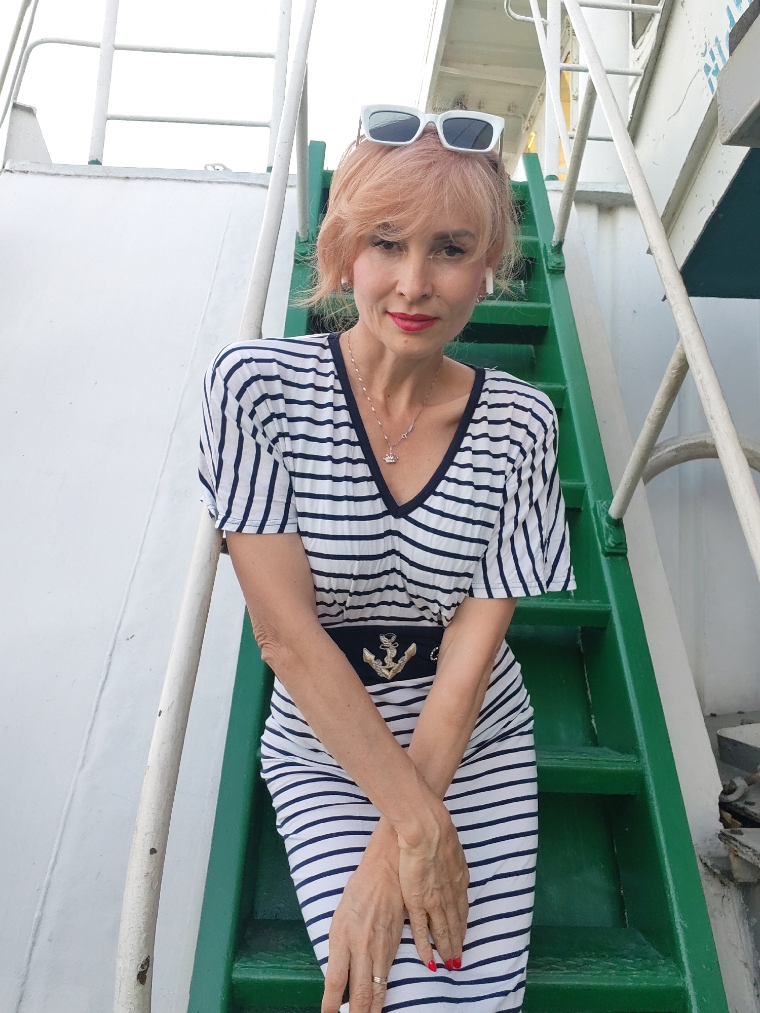 A woman with pinkish-blonde hair, wearing white sunglasses, a navy and white striped V-neck dress with a dark belt featuring an anchor emblem, and white earrings, stands on green steps. She has on red lipstick and a silver necklace. Her arms are crossed in front of her, and her hands are visible. She appears to be on the deck of a ship, suggested by the white railing and hull. The background is bright and overcast. The location is not specified to be in Shchetinino, Russia.