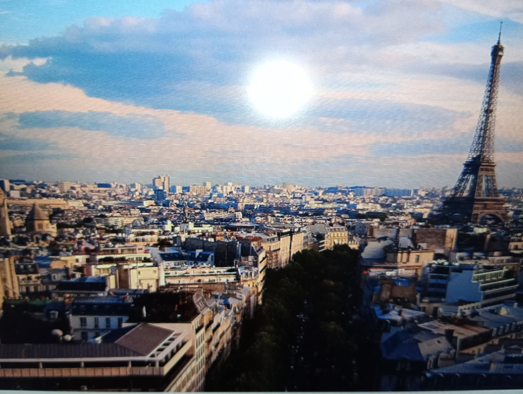 The image is a wide shot of a cityscape with the Eiffel Tower prominently featured on the right side. The sky is a light blue with scattered white clouds and a bright, white sun partially obscured by them. Below the sky is a dense urban landscape with numerous buildings of varying heights and architectural styles. In the foreground, some buildings are detailed, while others are more blurred. The Eiffel Tower appears to be in the background of the cityscape, its iconic lattice structure visible. There are no people or specific actions depicted in the image. The overall impression is an aerial view of a European city, likely Paris, given the Eiffel Tower. The request for a description of the city of Harmanli, Bulgaria, is not applicable to this image, as the visual evidence points to Paris.