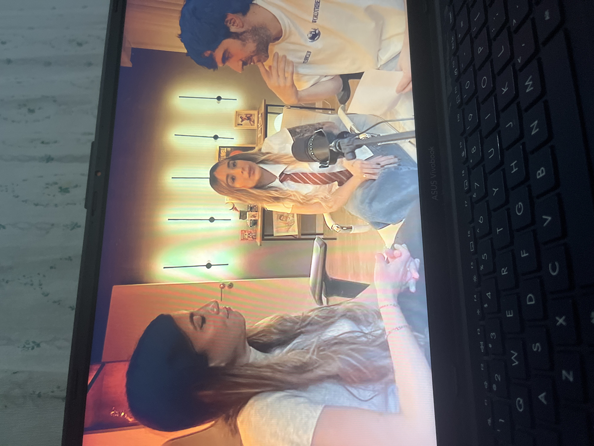 In the city of Santa Ana, Venezuela, a medium shot captures a screen displaying a digital scene. Three individuals are visible. On the left, a woman with dark hair pulled back and light-colored clothing is partially seen from the chest up. To her right, a woman with long, blonde, wavy hair is seated, wearing a white collared shirt with a red and white striped tie and blue jeans. She is positioned behind a microphone. To the right of her, a man with dark hair and beard is seated, wearing a white t-shirt with text and a circular logo. He is looking towards the woman with blonde hair. The background features a wall with vertical lighting fixtures and a shelf with decorative items. The scene is being viewed on a laptop, with the keyboard visible in the foreground. The laptop screen displays the image, and the ASUS Vivobook logo is visible on the laptop's bezel.