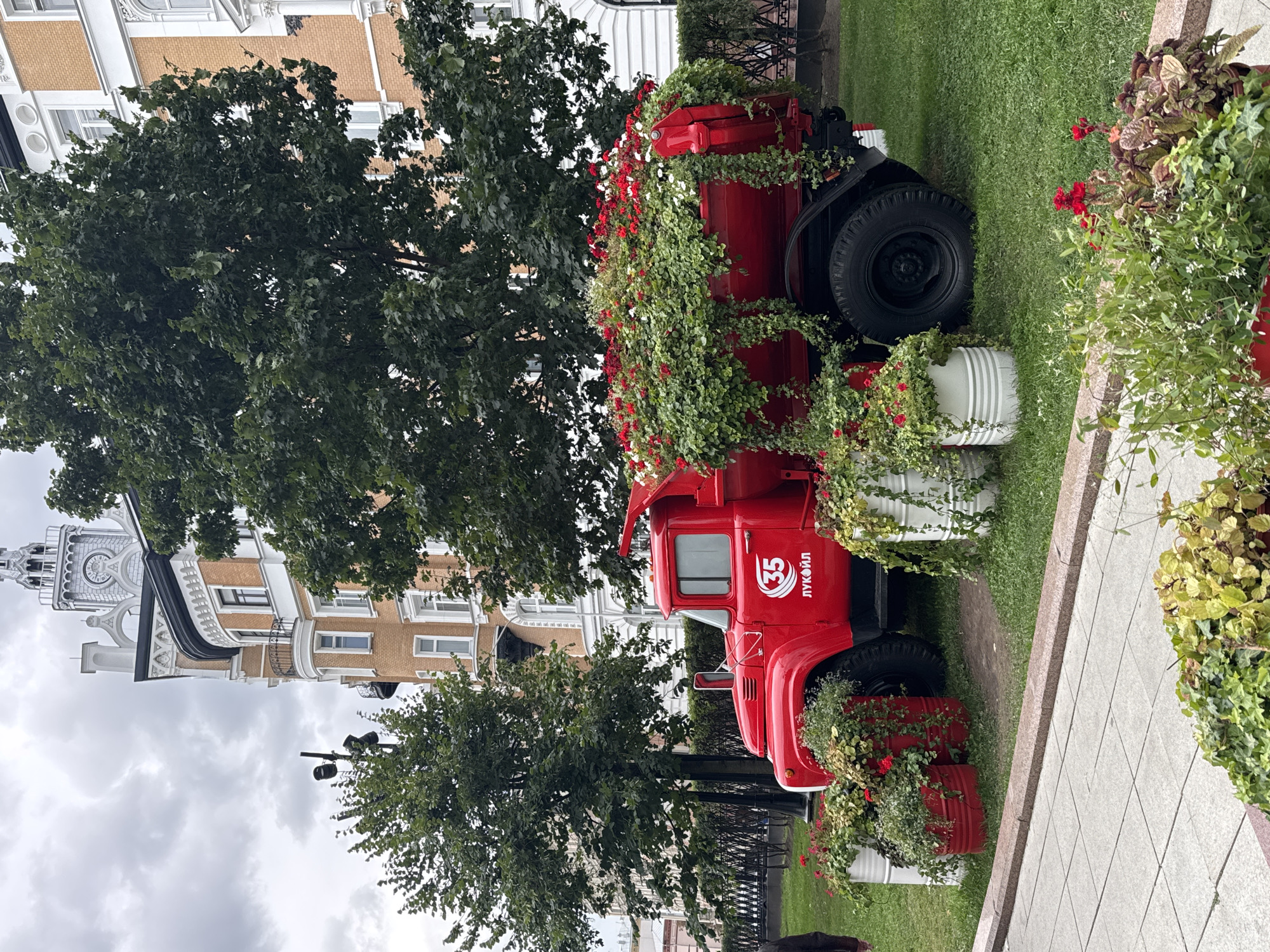 A bright red vintage truck, adorned with a profusion of red and white flowers, is parked on a grassy area in front of a grand, ornate building. The building, with its brick facade and intricate white detailing, features several windows and balconies, and is topped with a distinctive clock tower. Large green trees partially obscure the building. The truck's bed is overflowing with lush greenery and vibrant red and white flowers, creating a striking contrast with its bold red paint. Several white and red cylindrical planters, also filled with greenery and flowers, surround the truck. A paved walkway with a granite border is visible in the foreground, with more potted plants. The sky above is overcast, suggesting a cloudy day. The truck displays the number "35" within a circular emblem and the word "ЛУКОЙЛ" (LUKOIL) below it, indicating a possible advertising or promotional display for the Russian energy company. The scene appears to be set in a public space, likely a park or square, in the city of Moscow, Russia. There is one person, partially visible on the left side of the image, walking away from the truck.