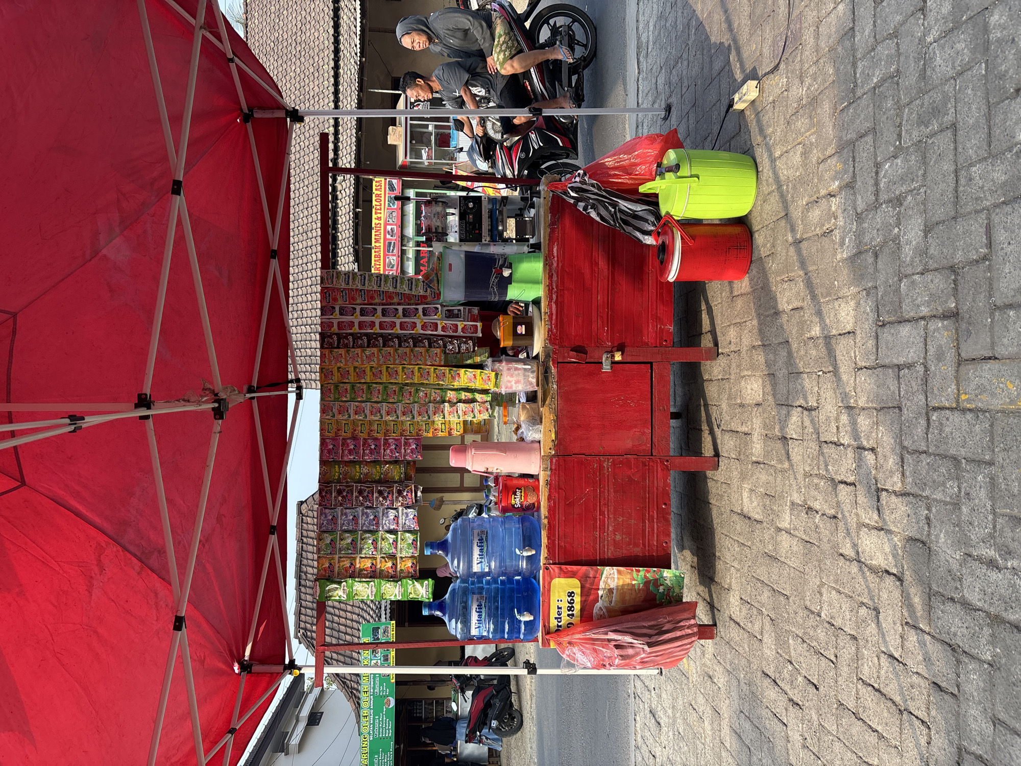 The image depicts a street vendor's stall in Tirtomoyo, Indonesia, under a red canopy. The stall is laden with various goods, including numerous packaged snacks and drinks displayed on the front. In the foreground, two large blue water dispenser jugs, labeled "Vitafit," are prominently placed. Next to them are a pink thermos and a red container labeled "Soklat." A red wooden cabinet serves as the base of the stall, with a yellow sign that reads "Order: 094868." Several red bags are hanging from the cabinet, and two large coolers, one red and one green, are positioned on the paved ground to the right. In the background, two people are seated on a motorcycle, facing left, with the stall between them and the street. The surrounding environment appears to be a street scene with buildings and a tiled roof visible behind the stall. The ground is paved with interlocking bricks, and shadows suggest it is daytime with the sun casting light from the right.