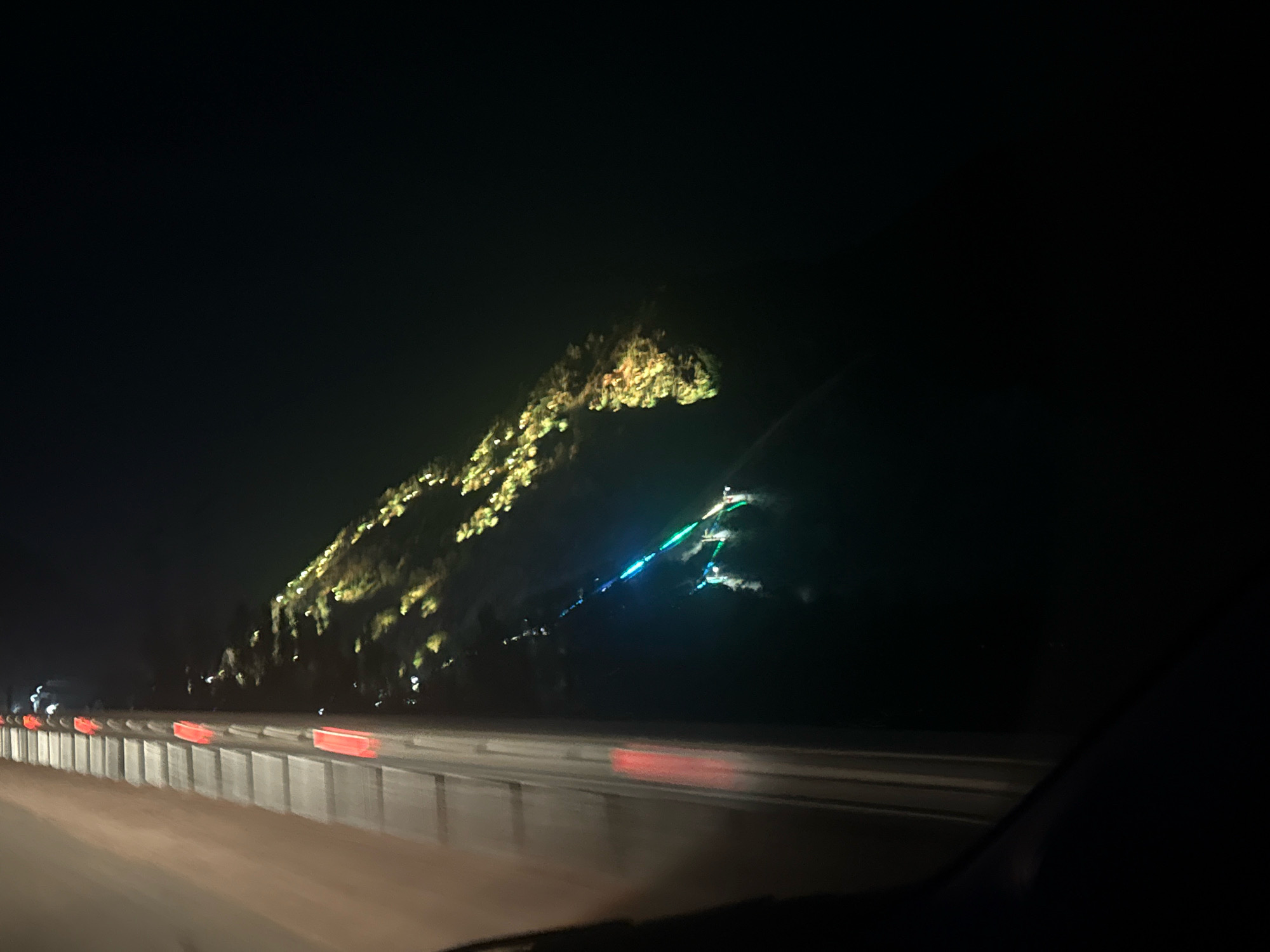 This is a night-time, motion-blurred photograph taken from a vehicle, likely on a highway. In the background, a large, dark hillside is illuminated by strings of yellow and white lights, creating a speckled effect across its surface. A section of the hillside features a multi-colored light display, with segments of blue and green lights visible. In the foreground, a concrete barrier with red reflectors runs along the side of the road. The road surface itself appears to be asphalt. The sky is completely dark, indicating it is nighttime. The location is identified as Arashan, Kyrgyzstan.