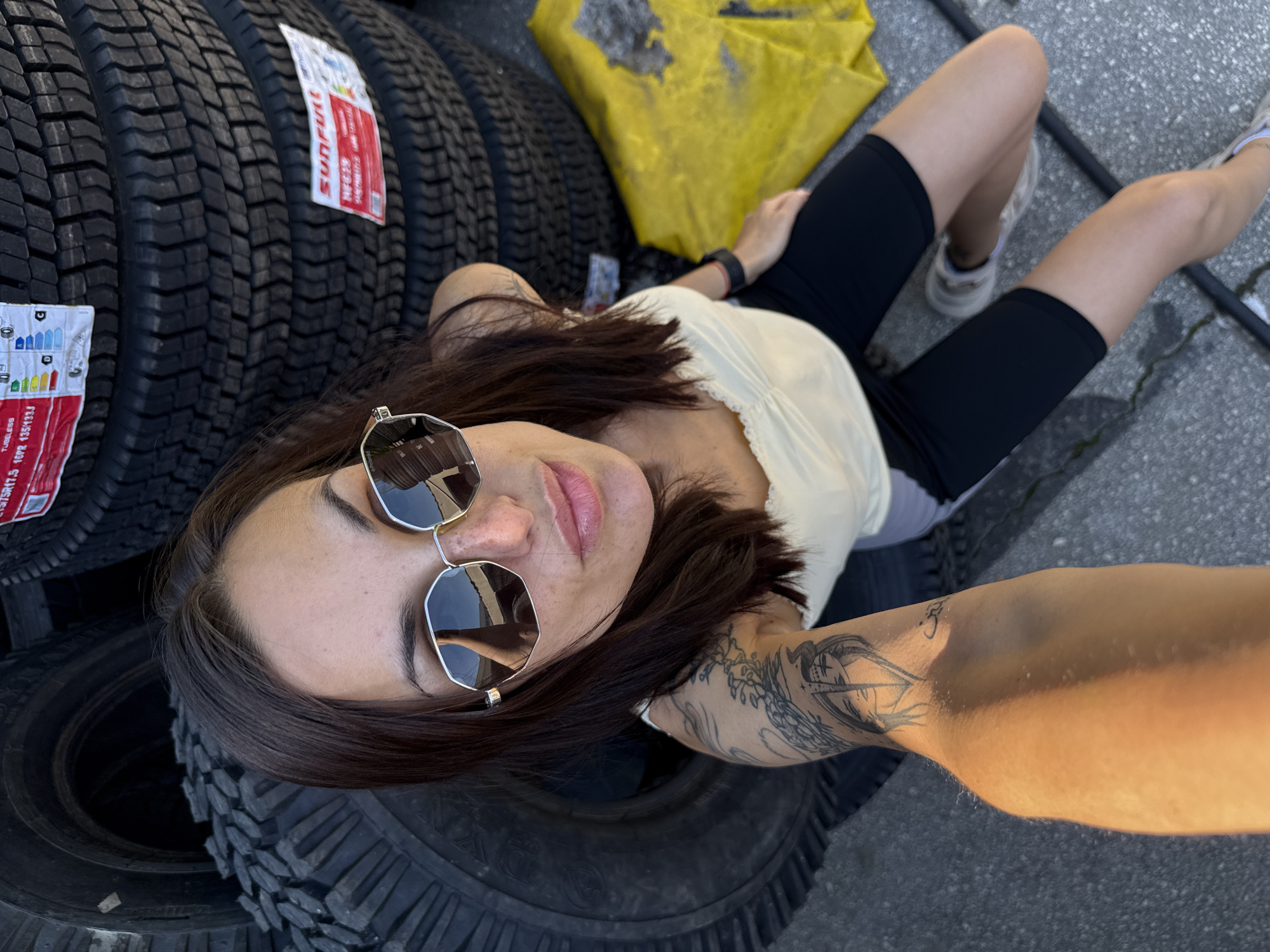 A woman with fair skin, dark brown hair, and wearing sunglasses is posing for a selfie. She is sitting amongst a stack of new tires. The tires have stickers with the word "Sunfull" on them and tire information labels. She is wearing a light-colored top and black shorts. She has tattoos on her left arm. She is sitting on a concrete surface with a yellow tarp or bag nearby. The image appears to be taken outdoors, possibly in a commercial area where tires are sold or stored. There is no information present in the image to support that the location is in Pleven, Bulgaria.
