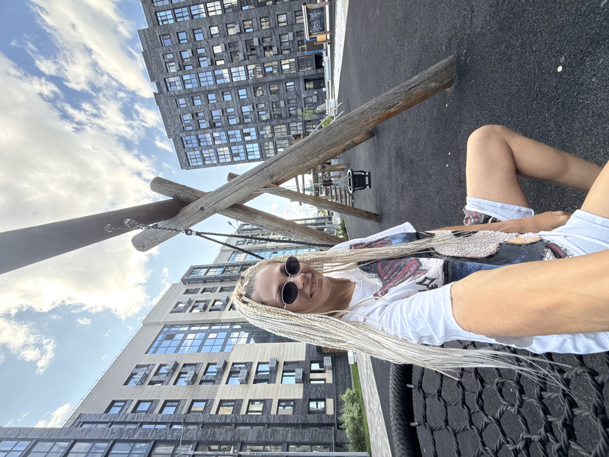 In Rusanovo, Russia, a young woman with blonde braided hair and oval sunglasses is seated on a black net swing. She is wearing a white t-shirt with a graphic print and light-colored shorts. The woman is holding a smartphone in her right hand, taking a selfie. Behind her, a wooden playground structure with a swing set and a baby swing is visible. In the background, modern apartment buildings rise against a sky filled with white clouds. The ground beneath the playground is a dark, rubberized surface.