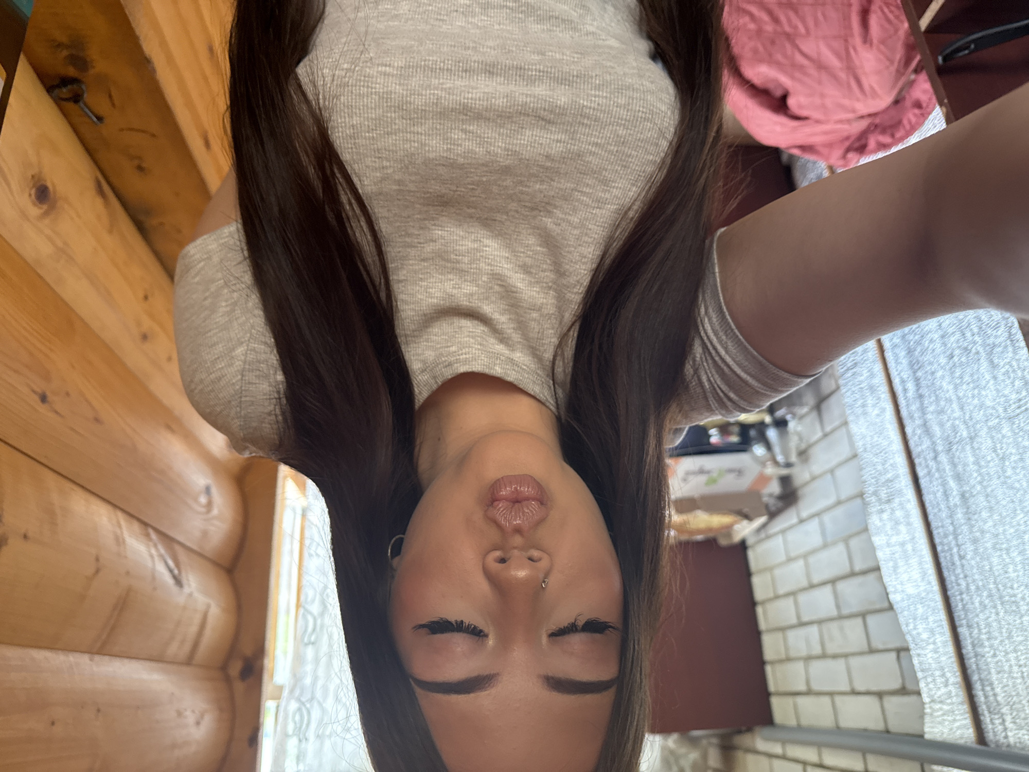 A woman is seen in a selfie, the camera oriented upside down. She has long, dark hair, closed eyes, and a visible nose piercing. She's wearing a light-colored, textured top. To the left, there's a log cabin-style wooden wall. In the background to the right, there is a pink garment and a white brick wall. This image does not provide enough information to confirm the location in the city of Belopakhotnyi, Russia.