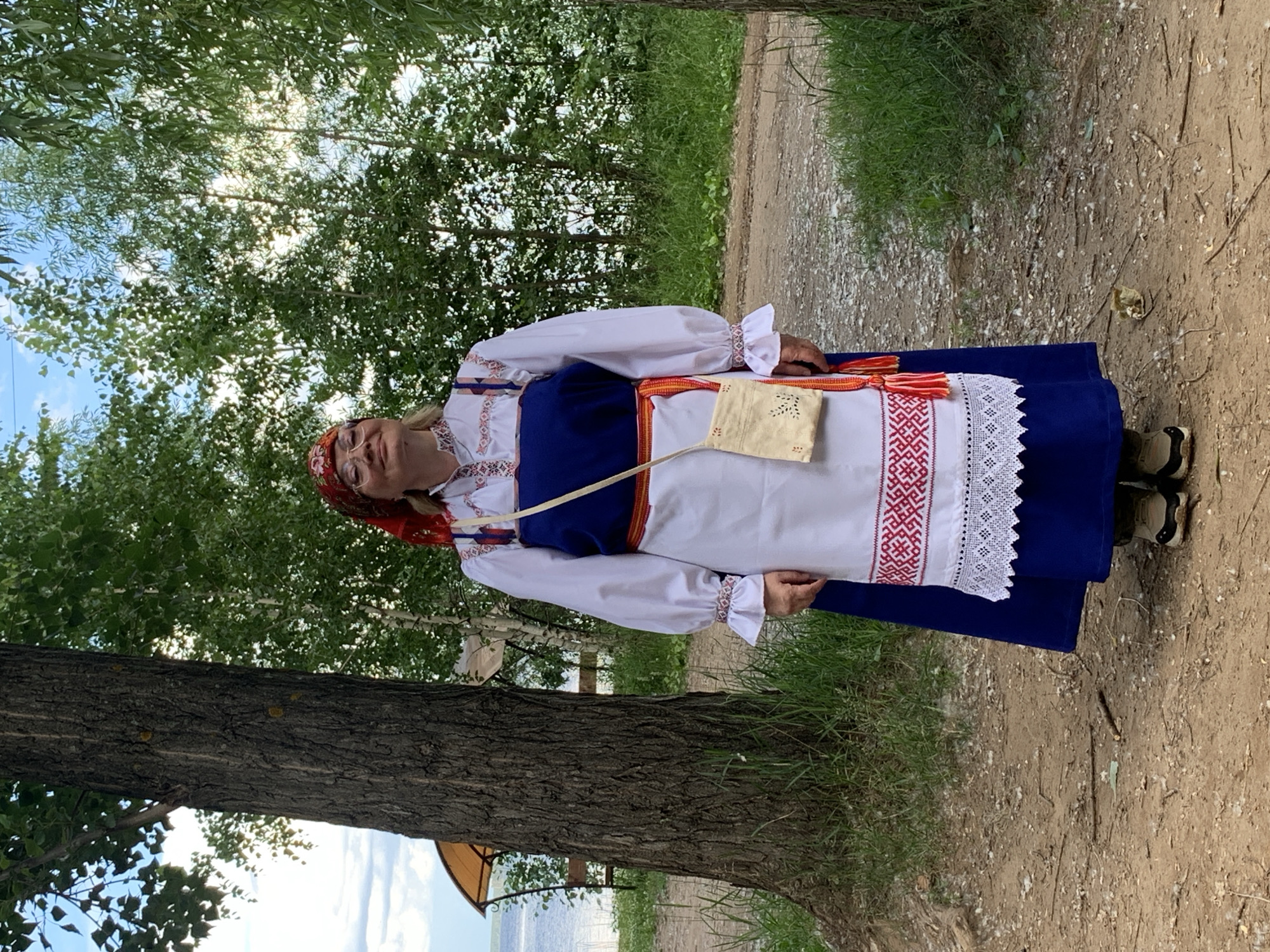 A woman, likely in her 50s or 60s, stands outdoors in what appears to be a rural or park-like setting. She is wearing traditional folk attire. Her outfit consists of a white blouse with long, full sleeves gathered at the wrists with ruffled cuffs, and a dark blue skirt. Over the blouse and skirt, she wears a white apron or overskirt with intricate red and white embroidery along the hem, and a decorative white lace trim. A red and white woven sash is tied around her waist, with tasseled ends. She also wears a red headscarf and glasses. A small, light-colored pouch with cut-out patterns is slung across her chest on a strap. She is smiling and looking towards the camera, with her head tilted slightly.
The background is natural, with lush green trees and foliage on the left side of the image. A dirt path runs diagonally across the scene from the upper right to the lower left. The ground is a mixture of dirt, small stones, and dried leaves. A large tree trunk is prominently visible in the foreground on the left. The lighting suggests it is daytime, likely late morning or afternoon, with dappled sunlight filtering through the trees. The weather appears to be pleasant and warm. The overall scene evokes a sense of traditional culture and a relaxed outdoor atmosphere. The woman's expression and attire suggest she might be participating in a cultural event or celebrating heritage. There is no visible text in the image.