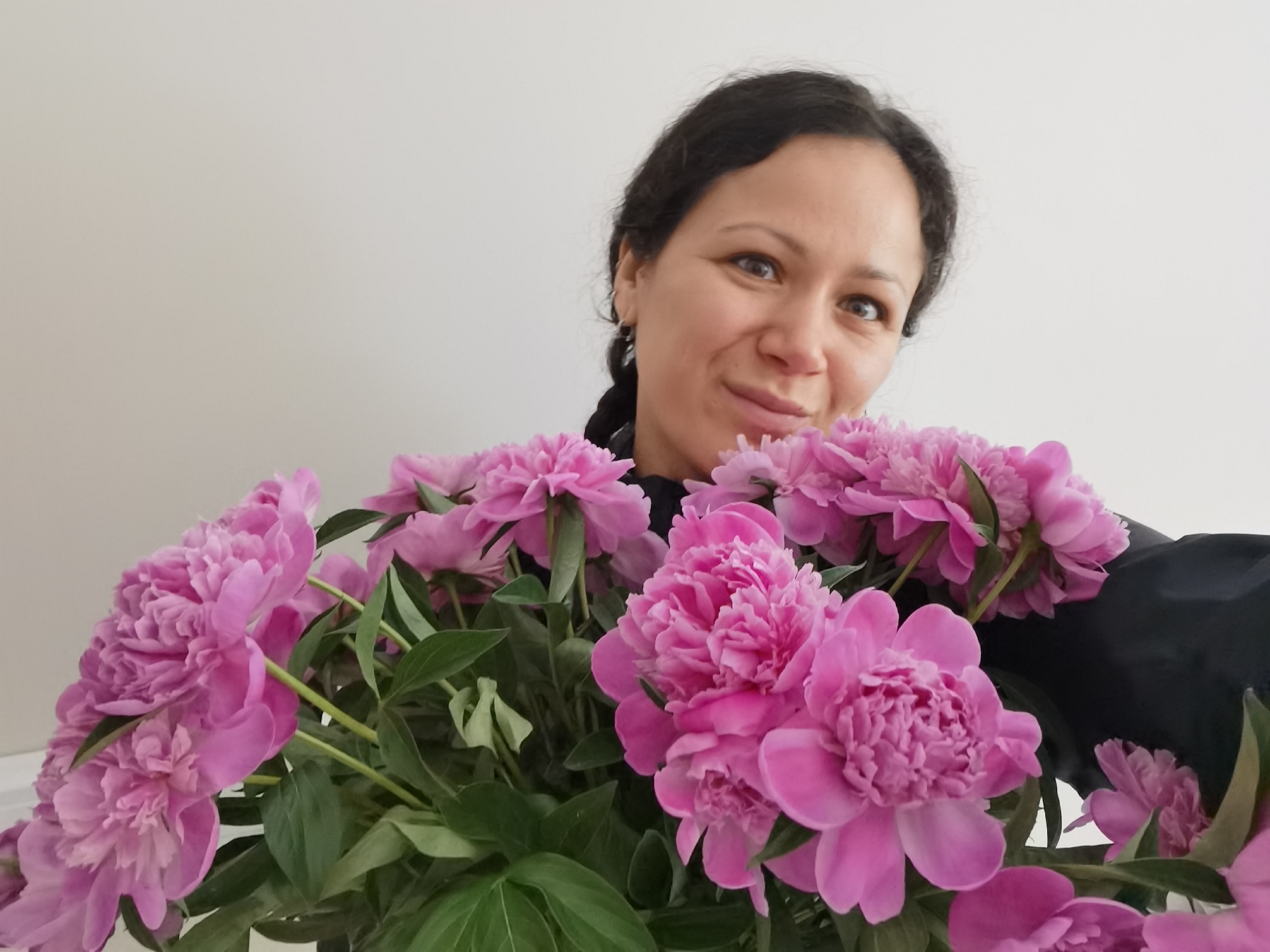 A woman with dark hair and a pleasant expression is captured in a medium close-up shot, looking directly at the viewer. She has dark, braided hair pulled over her left shoulder and brown eyes, and is wearing a dark-colored top. Her face shows a gentle smile, and she appears to be holding or standing behind a large, vibrant bouquet of pink peonies.
The peonies are in full bloom, showcasing numerous ruffled petals in various shades of pink, ranging from a lighter, softer pink to a more saturated fuchsia. Lush green leaves are visible among the flower heads, providing a contrast to the pink blooms. The bouquet is prominent in the foreground and mid-ground, largely obscuring the woman's body below her shoulders.
The setting appears to be indoors, against a plain, light-colored wall, possibly off-white or light beige, which creates a simple, uncluttered background that highlights the subject and the flowers. The lighting is bright and even, suggesting a daytime shot. There is no visible text in the image. The overall mood is cheerful and pleasant. The photo was taken in Chaikovsky, Russia.