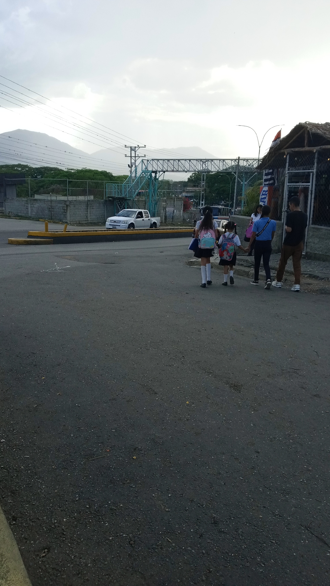 The image captures a street scene in an urban environment. In the middle ground, two young girls in school uniforms, wearing backpacks and white knee socks, walk hand-in-hand away from the viewer. To their right, an adult in a blue shirt walks alongside a person in a black shirt and brown pants.
Behind the group, a white pickup truck is parked next to a yellow curb, near a concrete fence. An elevated pedestrian walkway spans the scene in the background, with mountains visible in the distance. A small, shack-like structure with a thatched roof is located on the right side of the frame. The sky is overcast. The location is Unknown, Unknown.