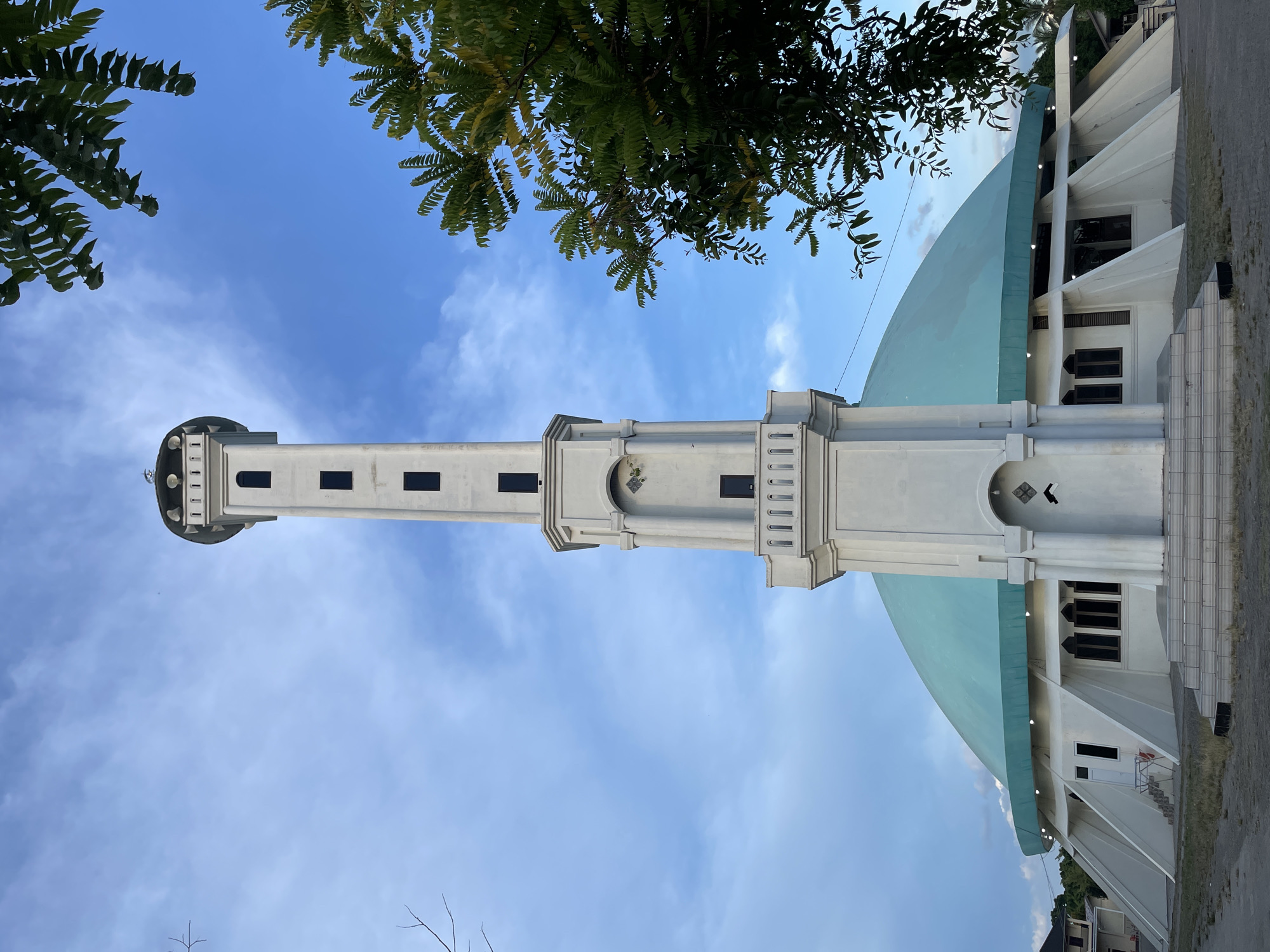 The image depicts the Masjid Raya Mujahidin mosque in Pasar Paloh, Pontianak City, West Kalimantan, Indonesia. The minaret and dome of the mosque are the central focus, positioned against a partly cloudy blue sky. Green foliage is visible in the upper corners of the frame. The building is white with a bluish-green dome. The mosque appears to be in an urban setting.