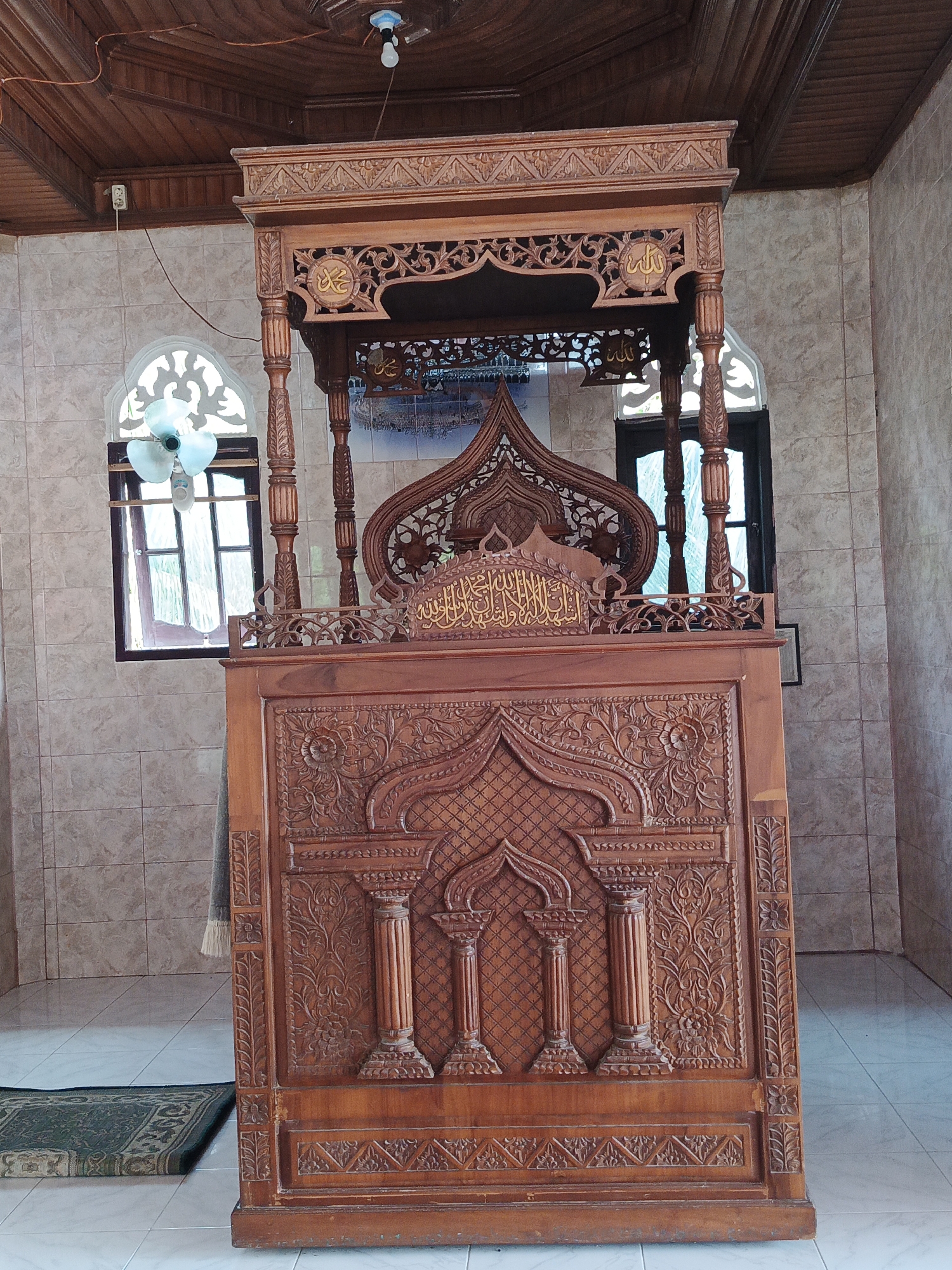 This media file captures a wooden minbar, a raised pulpit used in mosques for delivering sermons, located inside a room with tiled walls and a wooden ceiling. The minbar is elaborately carved with intricate patterns, including Arabic calligraphy and decorative motifs. The calligraphy on the minbar appears to be in gold.
The setting is a mosque or a prayer hall, suggested by the presence of the minbar and the overall architectural style, including arched windows. The light in the room appears to be artificial, with a light bulb hanging from the ceiling, and possibly natural light filtering through the windows. The windows have a decorative, arched top and are partially covered by thin curtains. A small, green prayer rug is visible on the tiled floor in front of the minbar. A ceiling fan is also present in one of the windows.
There are no people visible in the image. The scene is static, focusing on the detailed craftsmanship of the minbar. The time of day is not explicitly indicated, but the artificial lighting suggests it could be anytime, though the natural light through the windows hints at daytime. The weather is not discernible.
The location is identified as Sawang, Indonesia. The design of the minbar and the surrounding architecture may be characteristic of Indonesian mosque design. The overall impression is one of reverence and detailed artistic expression within a place of worship. The visible text is Arabic calligraphy on the minbar.