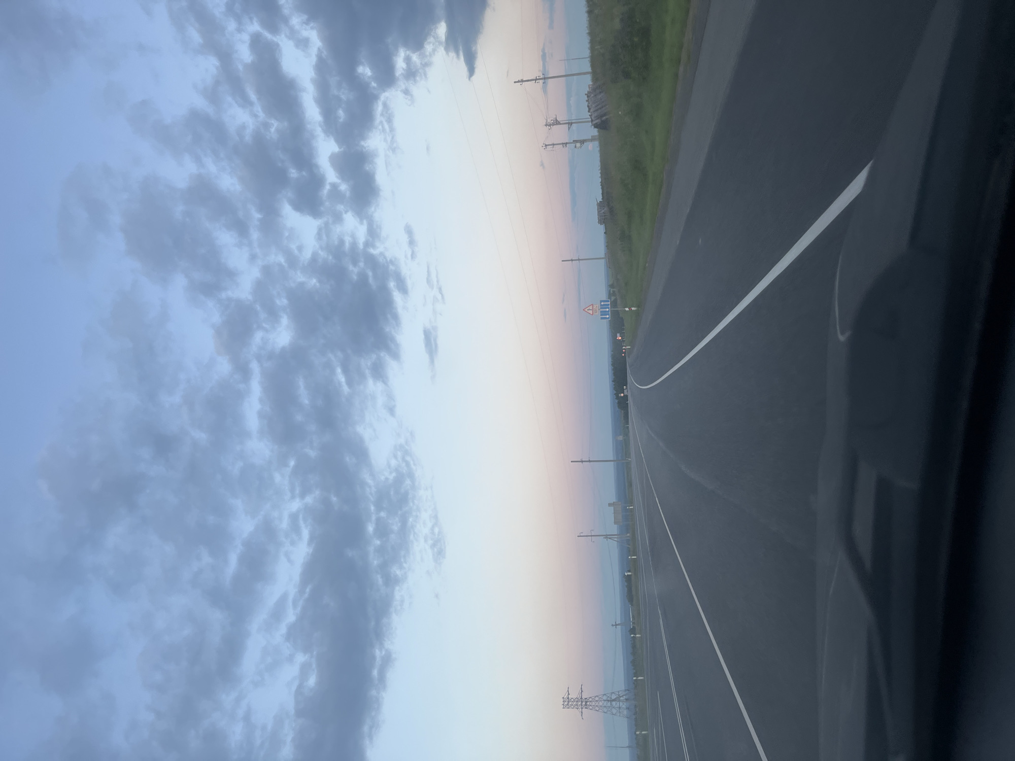 This media file shows a view from a vehicle traveling on a paved road in Tarasovo, Russia. The road appears to be a highway with a white dividing line and a shoulder. To the right of the road is a grassy embankment, and further in the distance are trees and power lines. The sky is visible, with dark clouds on the left and a gradient of light blue to pink towards the horizon. There are several electrical pylons visible along the side of the road, suggesting a rural or semi-rural setting. A road sign with a warning symbol is also visible. The time of day appears to be either sunrise or sunset, given the colors in the sky. The overall mood is calm and serene, with a sense of travel and open space.