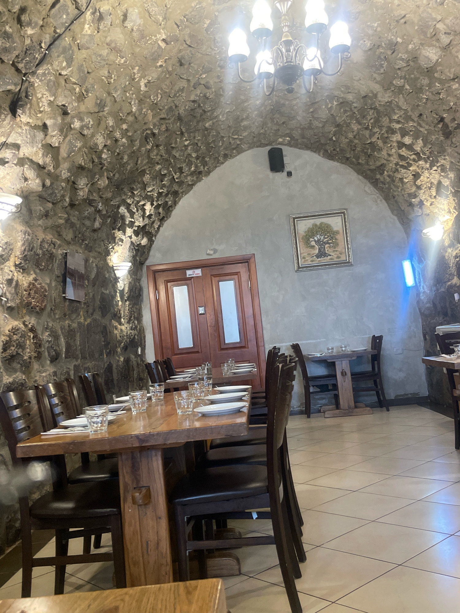 The image shows the interior of a restaurant in Tiberias, Israel. The restaurant features a rustic design with stone walls and a vaulted ceiling. Several wooden tables are set with plates and glasses, suggesting it is either set for customers or has recently served them. There are chairs around the tables, and some are empty while others appear occupied by blurred figures. A chandelier hangs from the ceiling, and there are wall lights illuminating the space. A framed picture of a tree hangs on a light grey wall. A wooden door with glass panels is visible in the background. The floor is tiled with a cream-colored pattern. The overall atmosphere appears to be dimly lit and cozy.