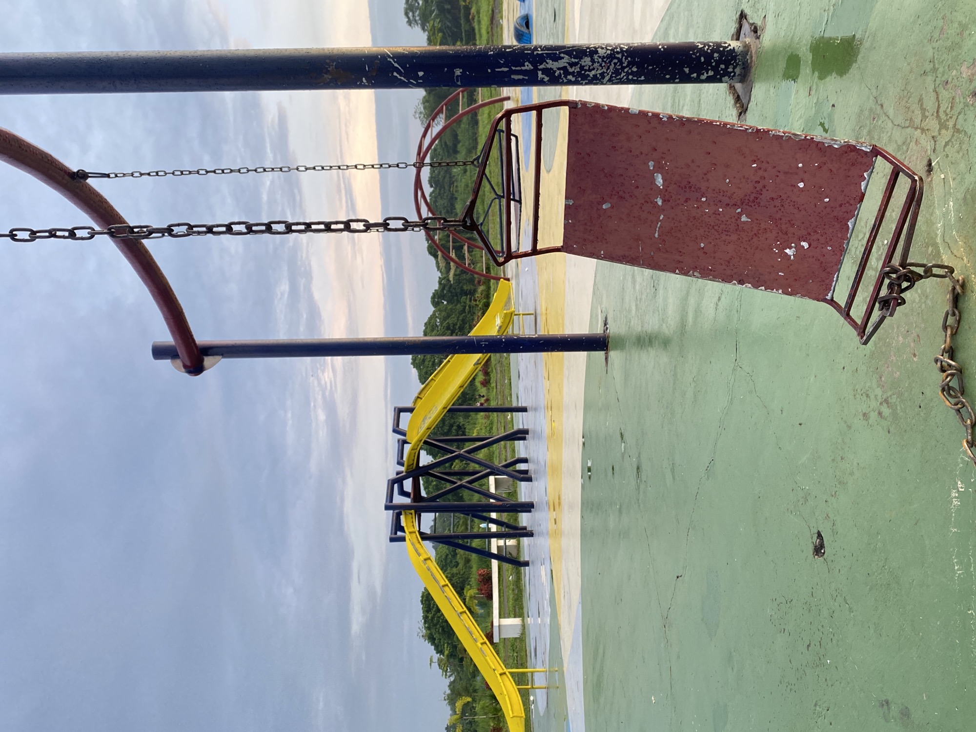 The image, captured in Davao City, Philippines, shows an outdoor playground under an overcast sky, suggesting either early morning or late afternoon light. The ground and equipment appear wet, indicating recent rain or high humidity.
In the foreground, a prominent reddish-brown metal swing seat, displaying significant rust and peeling paint, hangs from dark metal chains. These chains are attached to a dark blue/black metal support frame, which also exhibits considerable paint chipping and wear. Another set of chains hangs to the left, without a visible seat. The ground beneath the play equipment is a hard surface, painted in faded sections of green and yellow, marked by visible cracks, dirt, and wet patches.
In the mid-ground, a bright yellow slide with a winding structure is visible, supported by dark metal frames similar in style and condition to the swing set. Beyond the playground structures, a dense backdrop of lush green trees and foliage extends towards the horizon, framed by the soft, diffused light of the cloudy sky.
There are no people present in the scene, and no visible text is discernible on any of the objects or surfaces. The overall impression is one of a well-used, somewhat weathered public recreational area.
