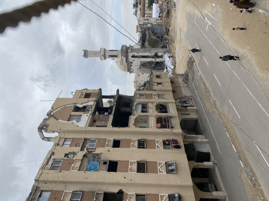 The image displays a street scene in Khan Yunis, Palestinian Territory, characterized by significant structural damage. In the foreground, a multi-story, beige-colored building with brown decorative elements stands prominently on the left. This building exhibits extensive damage, including large sections of missing exterior walls revealing internal structures, shattered windows, and numerous impact marks across its facade. A blue tarp covers a damaged window opening on an upper floor, and laundry hangs from balconies on lower floors. Arched alcoves are visible on the ground level, with a small white car parked beneath one.
An asphalt road occupies the midground, featuring white double solid lines and a directional arrow. Two adult males walk on the road, one closer to the camera and one further away. On the right side of the road, a group consisting of an adult and several children stands on a dirt shoulder.
In the background, a heavily damaged white mosque with a distinct dome and a tall minaret is visible. Large sections of the mosque's lower structure are collapsed into rubble. The area between the foreground building and the mosque is largely cleared, exposing an expanse of debris and construction remnants, including some temporary coverings. The sky is overcast.