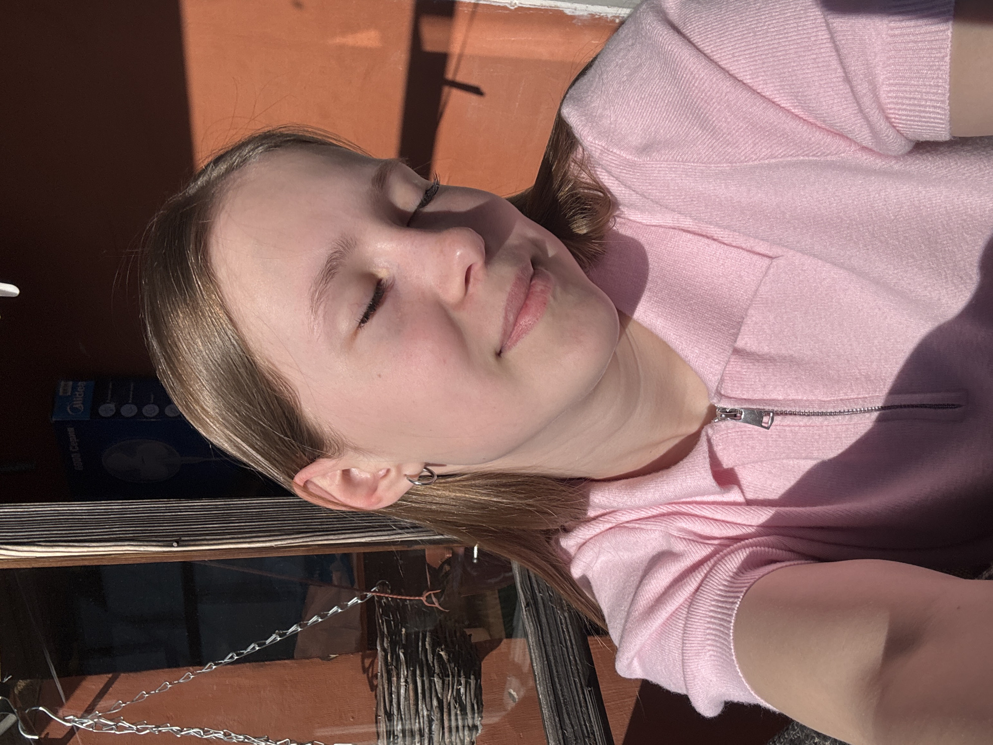 A young person, likely female, with light brown hair, is captured in a close-up, upward-angled shot, with their eyes closed and face turned towards a bright light source, presumably the sun. They appear to be basking or resting in direct sunlight, which casts strong, defined shadows across the scene. The individual wears a light pink, long-sleeved top made of a ribbed or knitted fabric, featuring a silver zipper detail at the neckline. A small, thin silver hoop earring is visible in their left ear.
The setting appears to be outdoors or semi-outdoors, possibly on a balcony or patio in Chaikovsky, Russia. The background features an orangey-brown wall or floor surface, with distinct shadows from unseen structures or objects. In the lower left of the image, metal chains are visible, suggesting a railing, fence, or hanging decoration. Part of a dark blue box or packaging is visible behind the person's head, with the brand "Midea" and the Russian word "Фан" (Fan) partially legible on it, suggesting an appliance box. The bright, direct sunlight indicates daytime, likely a clear and sunny day. The overall mood conveyed is one of peace and relaxation.