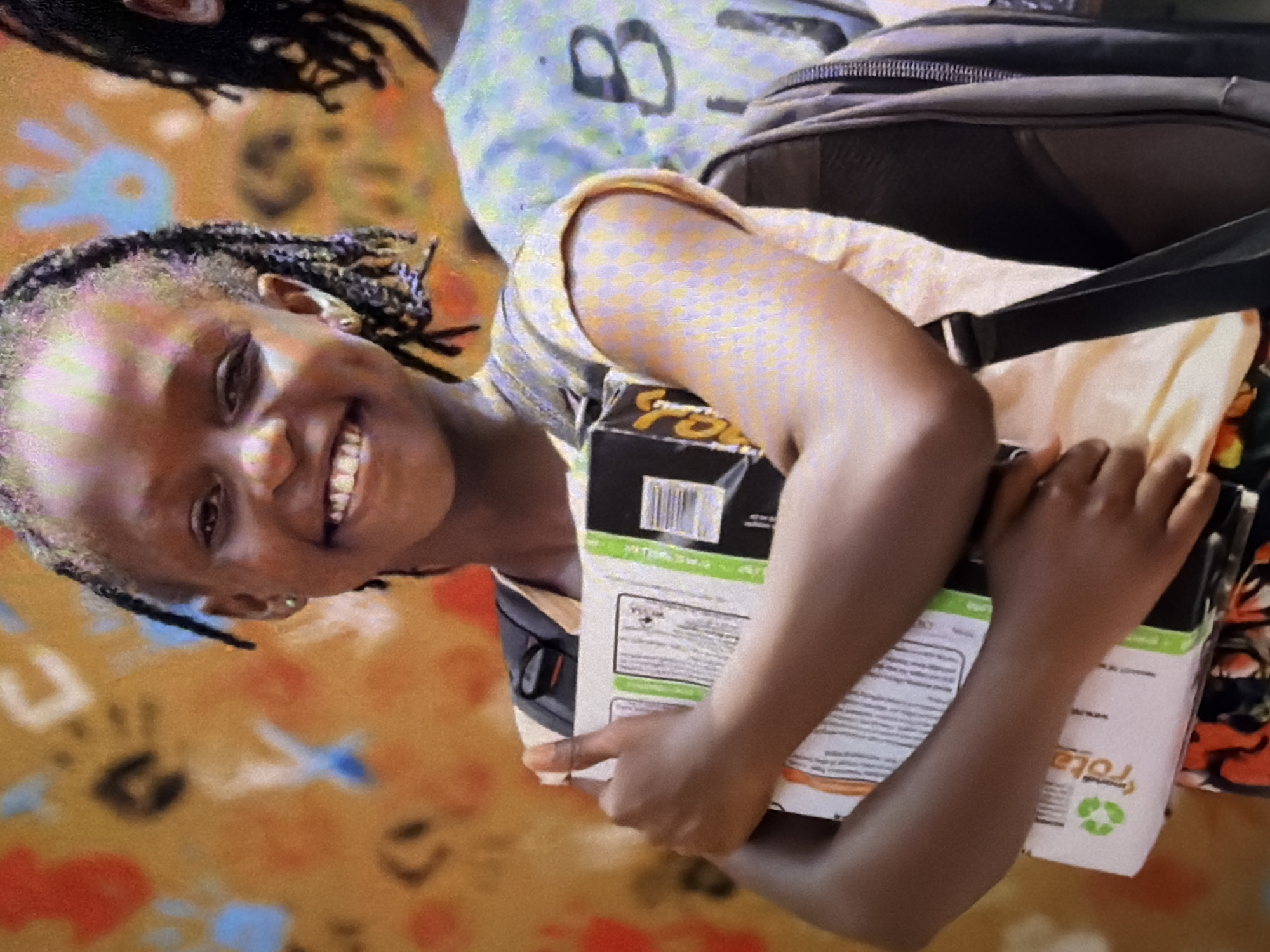 A young Black girl, with braided hair and a bright smile, is the focus of the image. She is wearing a light-colored, possibly yellow, short-sleeved shirt with a subtle patterned texture. Her left arm is wrapped around a cardboard box or book, which she is holding against her chest. A portion of a backpack is visible on her right side, with dark straps. Her lower body is partially visible, wearing a colorful patterned skirt. She has a small gold earring in her left ear. Behind her, a wall with colorful handprints and abstract shapes serves as the backdrop. The scene appears to be outdoors or in a well-lit indoor space, given the vibrancy of the colors and the natural lighting. To the right of the girl, another person, also Black and with braided hair, is partially visible, wearing a light-colored t-shirt with a large black "B" printed on it. The overall impression is one of joy and perhaps a school or community setting.