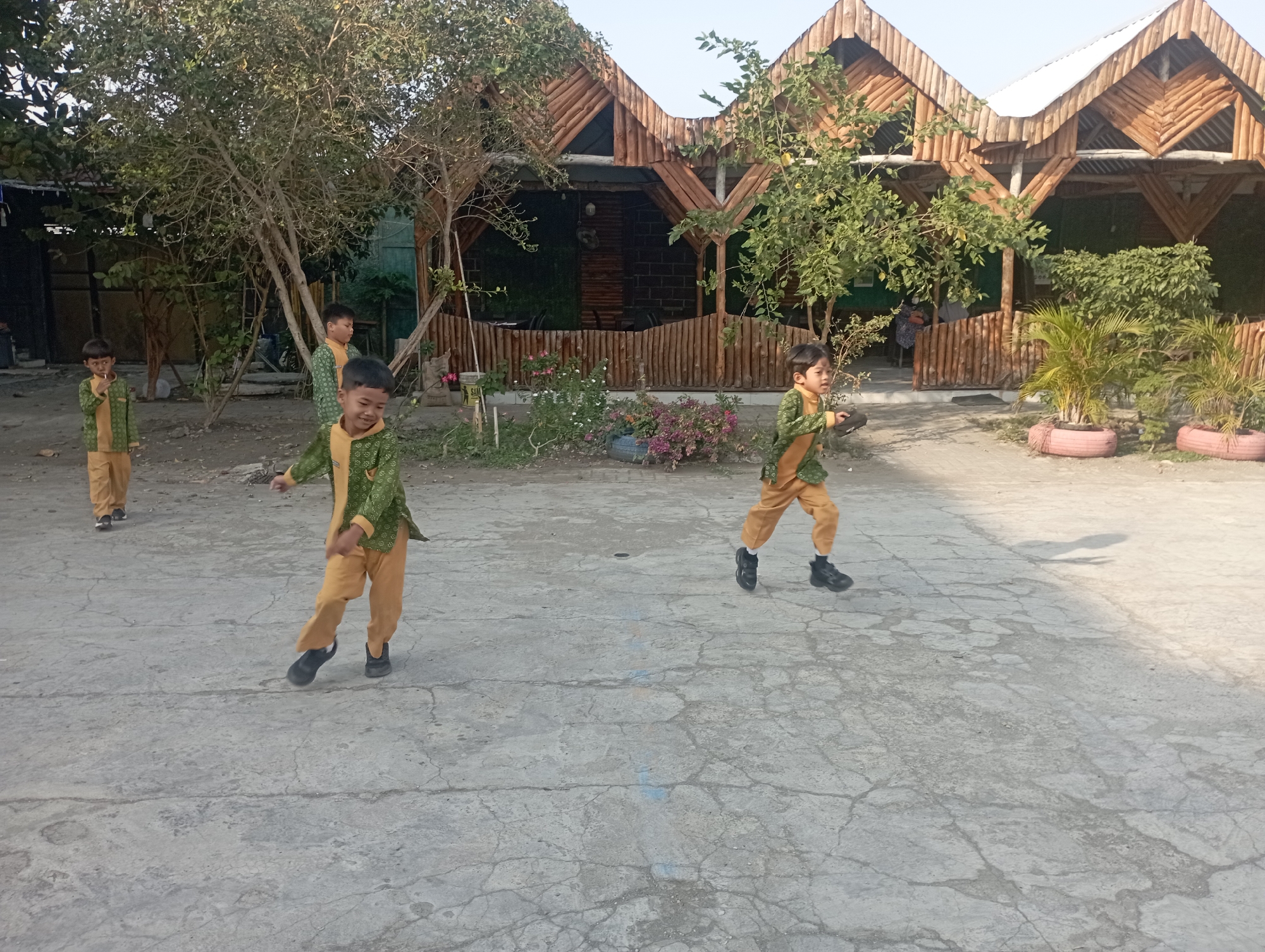 In Kramat, Indonesia, four young boys are captured in a moment of outdoor activity. Three of the boys are prominently featured in the foreground, dressed in matching light green tunics with gold accents and tan pants. The boy on the far left is standing, holding a light-colored object to his mouth. The boy in the center is in mid-stride, his arms outstretched as if running or playing. The boy on the far right is also in motion, his right leg raised. A fourth boy, partially obscured by foliage, stands further back on the left, also in similar attire.
The setting appears to be an open, paved area with a textured, concrete surface marked by cracks. Behind the boys, a rustic, multi-tiered wooden structure with a distinctive zigzag roofline dominates the background, suggesting it might be a restaurant or recreational facility. Lush green trees and bushes are scattered throughout the scene, with flowering plants in shades of pink and purple visible near the wooden building. Two large planters made from repurposed tires hold potted palm plants. The sky above is a pale, hazy blue, indicating it's daytime. The overall impression is of a casual, sunny day in a semi-developed outdoor space.