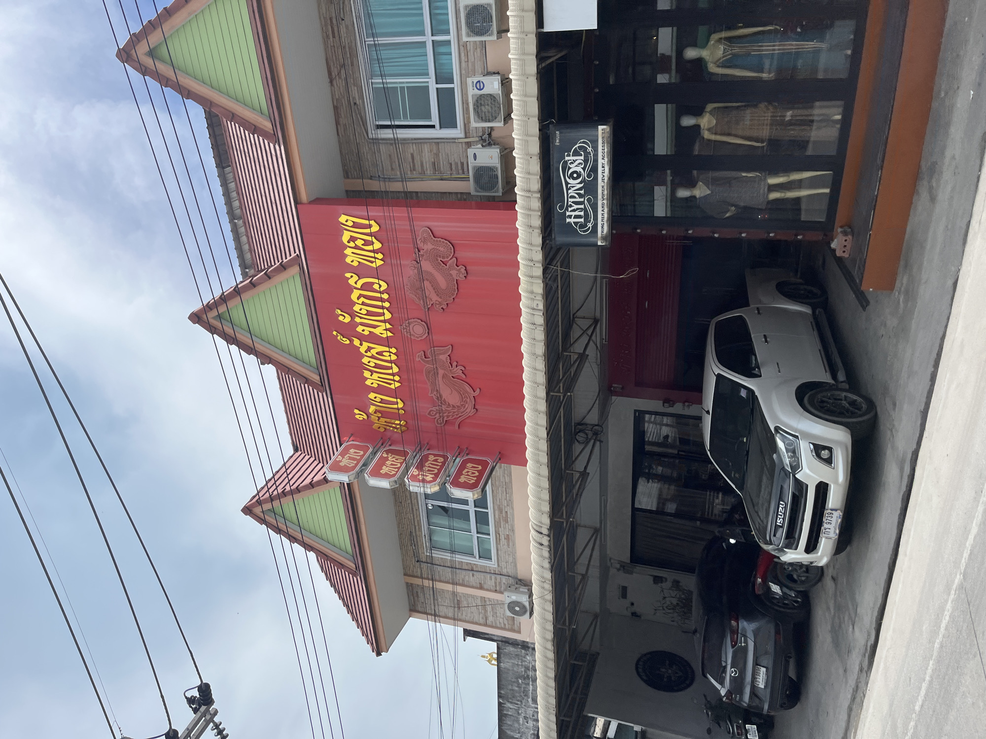 The image shows a street view in Koh Phangan, Thailand, featuring a building with a red facade and yellow Thai lettering. The building has a prominent pitched roof with green gable ends and red tiles. Below the upper story, a covered walkway extends outwards, supported by metal poles and a white awning. This area houses parked vehicles, including a white Isuzu pickup truck with a black hood and a dark-colored SUV. To the right, a storefront with mannequins displayed inside is visible, with a black sign that reads "HYPNOSE" and "CLOTHING MEN AND WOMEN JEWELRY ACCESSORIES". Electrical wires are strung across the sky above the building. The environment appears to be an urban or commercial area.