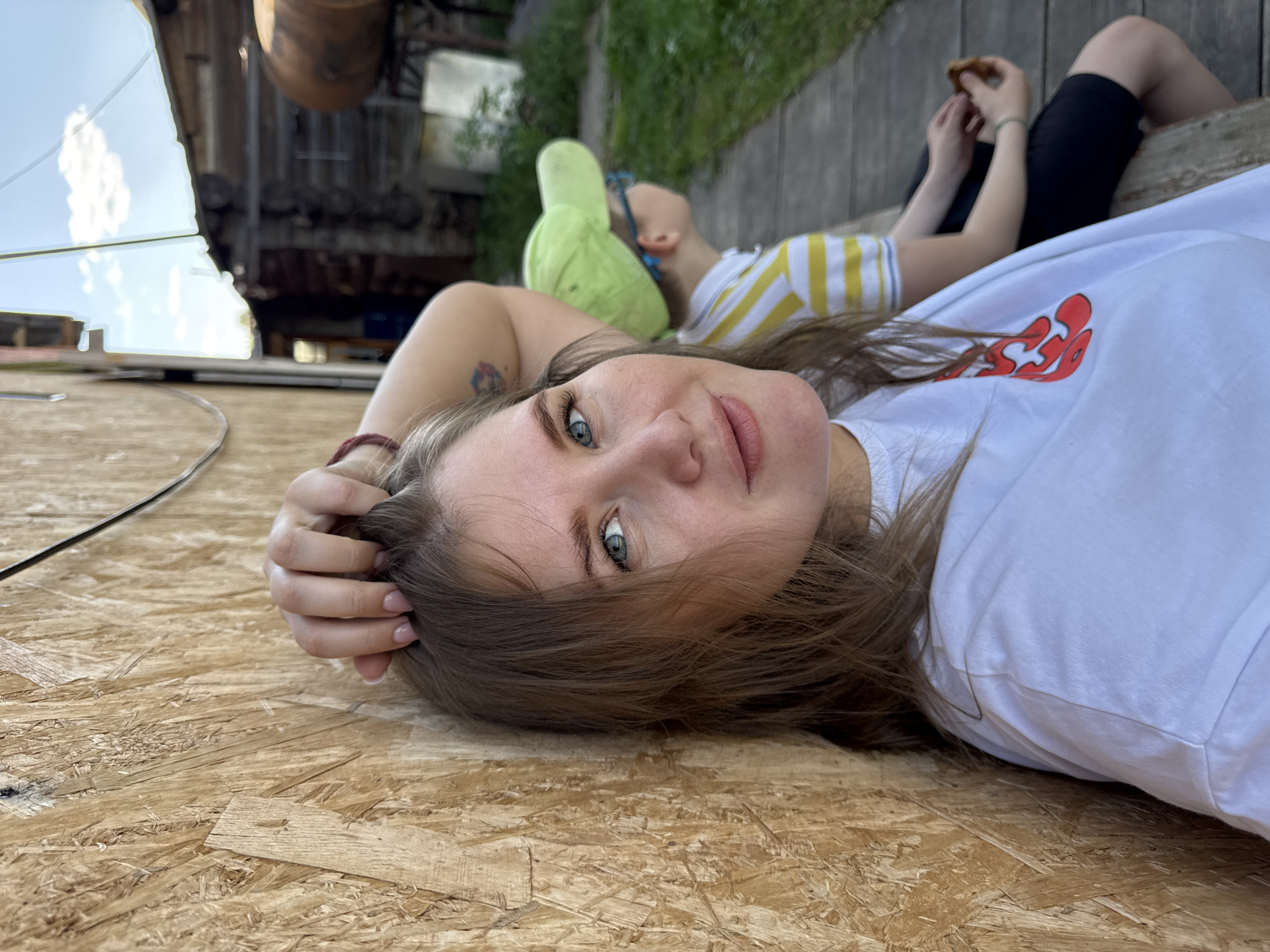 The image depicts a woman lying on what appears to be a wooden or plywood surface, possibly a deck or platform. She is wearing a white t-shirt with a red design and has brown hair and light-colored eyes. Her head is resting on the surface, and her hand is holding her hair. In the background, there is a child wearing a yellow and white striped shirt and a green hat. They are lying on a wooden surface that looks like a raised deck or platform. Behind the child, there is another person eating a snack and wearing black shorts. In the far background, there is some industrial-looking metal construction, and foliage can be seen. Based on the location information (Фоки, Russia), it is possibly a recreation area or park with industrial art installations.