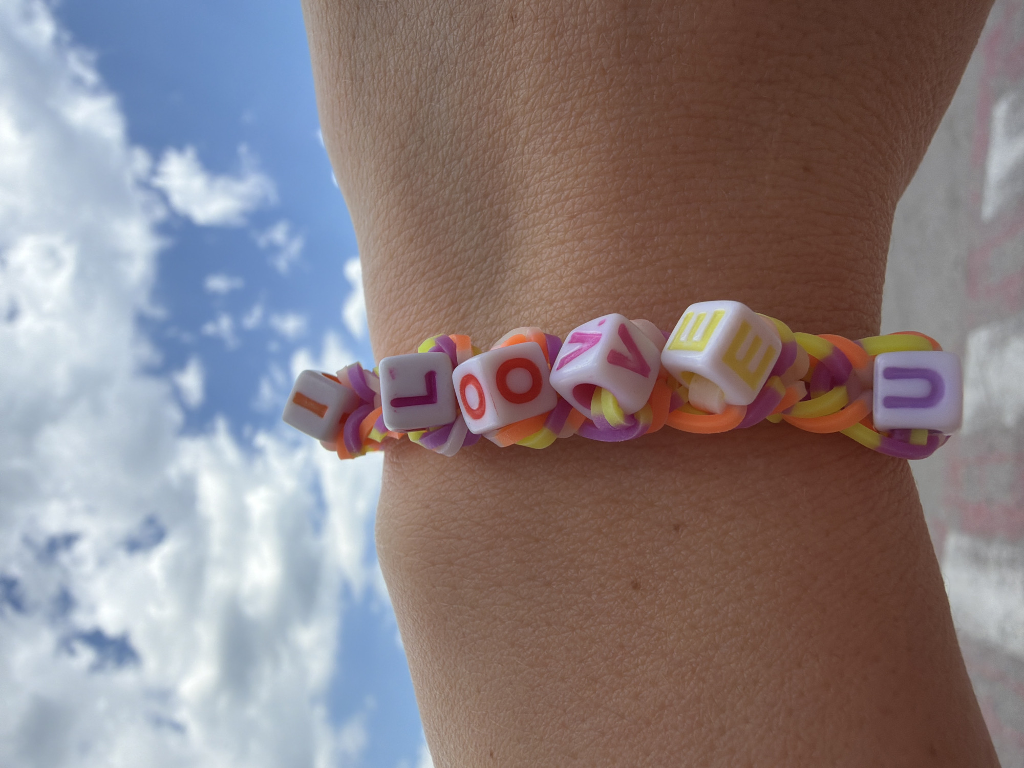 The image shows a person's wrist wearing a handmade bracelet spelling out "LOVE" with letter beads strung onto colorful rubber bands. The bracelet is positioned against a backdrop of a partly cloudy sky. The arm appears to be light-skinned. The scene is outdoors, likely in a well-lit area. No specific identifying landmarks indicate the location within Budva, Montenegro.