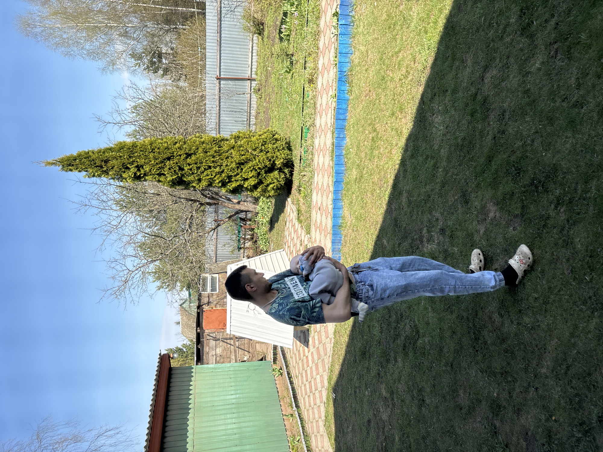 A man is holding a baby in a grassy yard in Мещёры, Russia. The man is wearing jeans, a t-shirt, and Crocs. The baby is bundled in grey clothes. The yard has a brick path with a blue painted border. Fences and a small shed are visible in the background, along with trees. The scene is brightly lit.