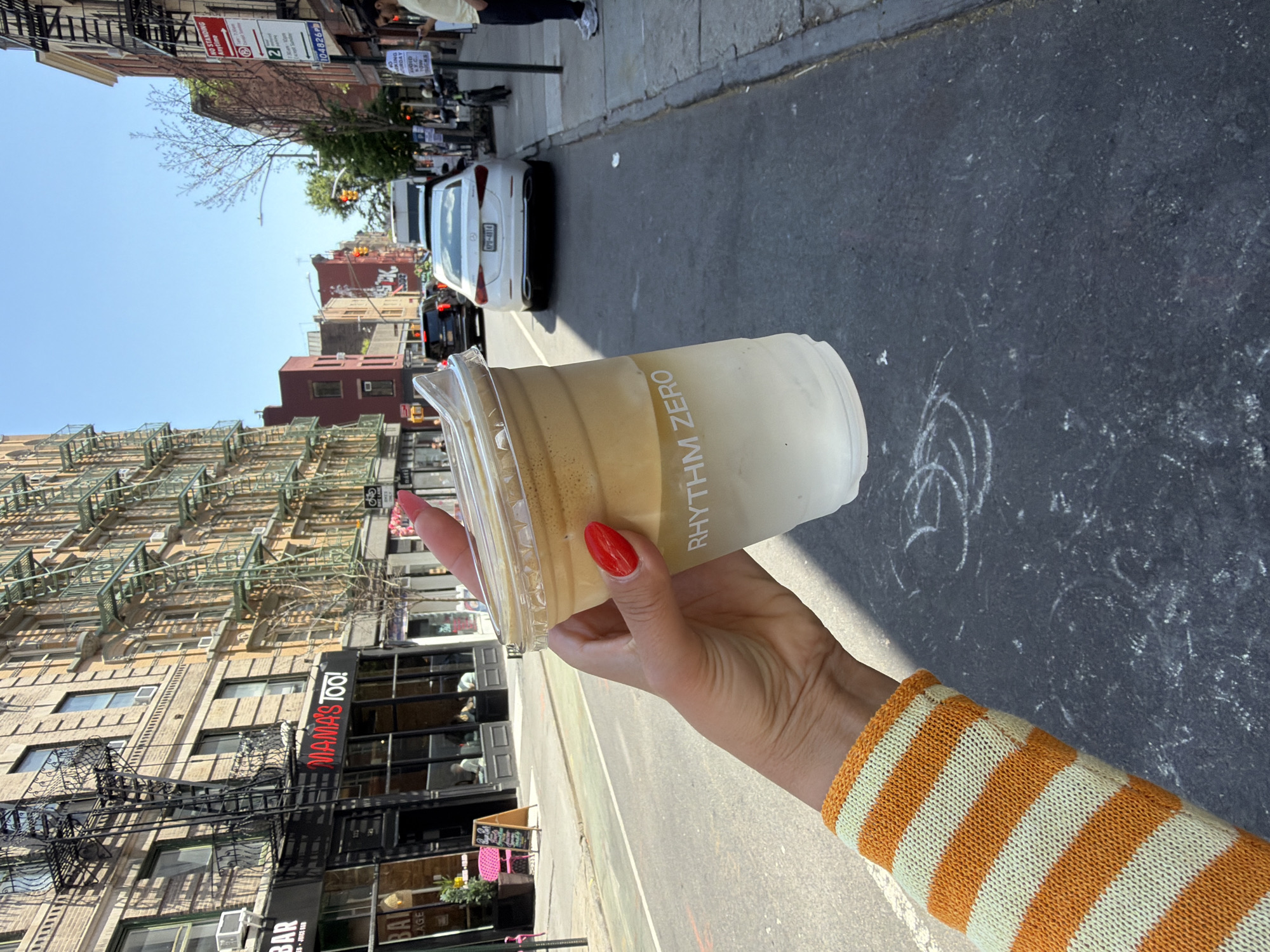 A vertical, medium-shot captures an arm and hand holding an iced coffee cup against the backdrop of a New York City street. The hand, with red nail polish, firmly grips the cup, which is labeled "RHYTHM ZERO". The arm is clad in a striped orange and white sleeve. The street scene includes a white car, various buildings, and architectural details like fire escapes. A restaurant sign is visible in the background with lettering "MAMA'S TOO!" in dark letters. Road markings and vehicles are also present, suggesting a typical urban environment. Street signs are present as well, containing the letters "KING".
