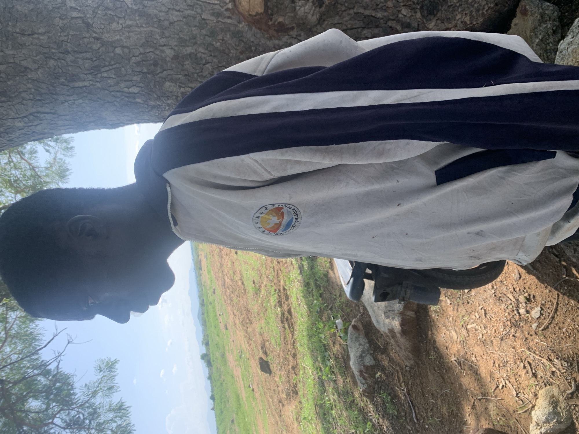 The media file shows a young person from the side, with their head tilted down. They are wearing a white jacket with dark blue stripes. A circular emblem with Chinese characters and an image of birds and a sun is visible on the jacket. The person appears to be sitting next to a tree with rough bark. In the background, there is a field with dry, brown soil and patches of green grass, suggesting a rural or agricultural setting. The sky is visible and appears bright, indicating it is daytime. A portion of a vehicle, likely a motorcycle or scooter, is visible in the lower right portion of the image, partially covered by a light-colored tarp. The person's profile is silhouetted against the bright sky, making their facial features unclear. The overall impression is of a casual outdoor scene in a rural environment in Nigeria.
