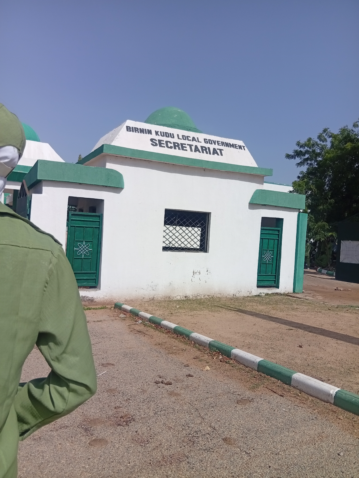 This is an outdoor, eye-level photograph of the Birnin Kudu Local Government Secretariat building in Nigeria, taken on a clear, sunny day. The building is painted white with prominent green trim and features several green, dome-shaped roofs, two of which are visible.
The words "BIRNIN KUDU LOCAL GOVERNMENT" and "SECRETARIAT" are boldly displayed in dark letters on the front facade of the building. The building has multiple green metal doors with decorative patterns and windows fitted with dark security grates.
In the left foreground, a person is partially visible from the back and left shoulder, wearing a military-green uniform or work attire. The person's presence suggests activity around the secretariat.
The ground in front of the building is a mix of gravel and dirt. A low curb, painted in alternating sections of green and white, runs along the edge of the paved or gravel area. Green trees are visible in the background on the right side under a bright, clear blue sky, indicating daytime with good weather. The overall scene depicts a functional government facility in a sunlit, possibly semi-arid, environment.