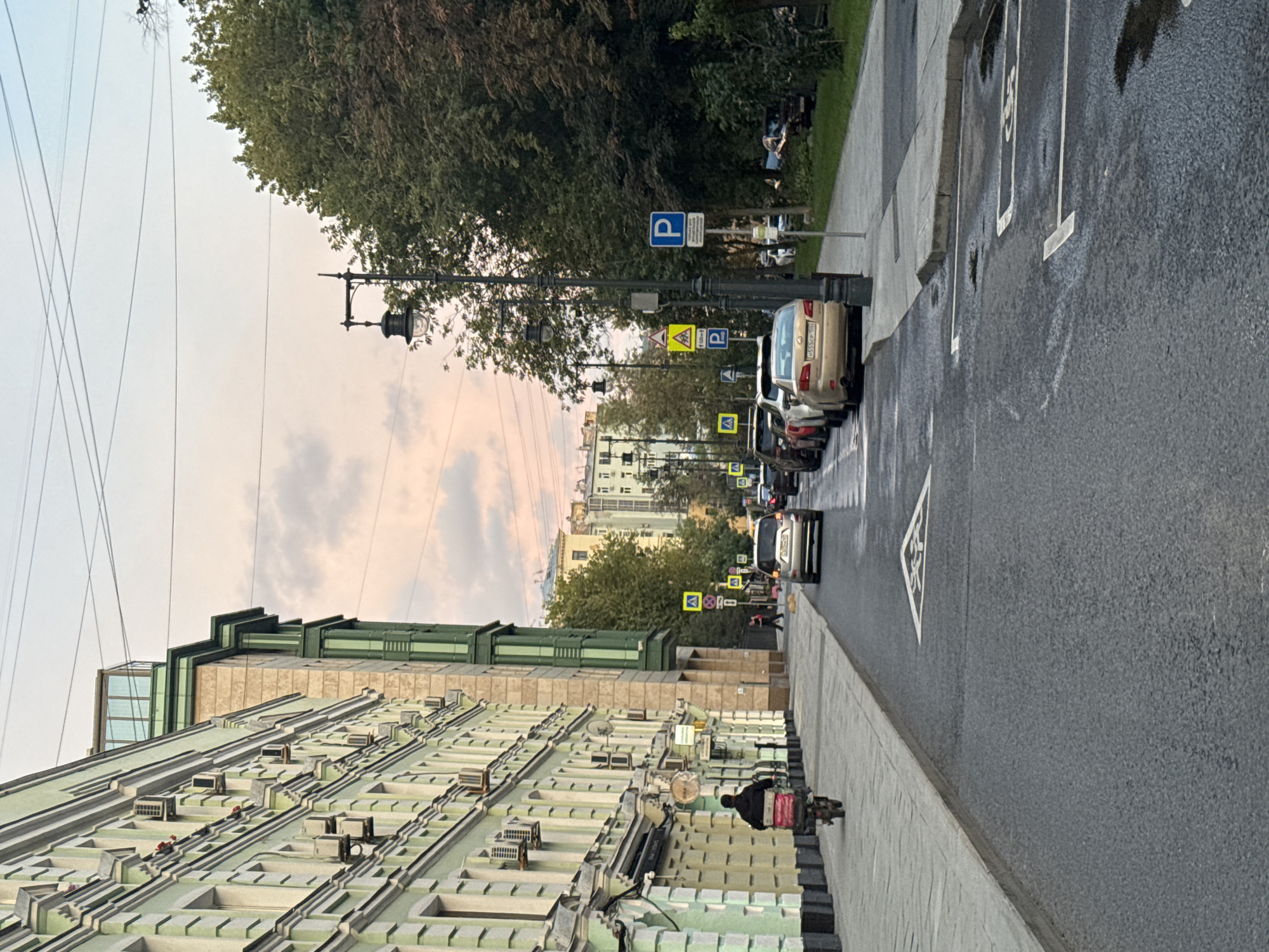 The image captures a street scene in Moscow, Russia, likely during late afternoon or early evening, given the soft, diffused lighting and the pastel hues in the sky. A pale green, multi-story building with numerous air conditioning units lines the left side of the street. A person on a scooter with a red and white cargo box is traveling away from the camera on the sidewalk.
The street itself is paved and shows signs of recent rain with wet patches and reflections. Several cars are parked along the right side of the street, and others are in motion. A prominent street lamp with a decorative lantern stands on the right. Various traffic signs, including parking signs (indicated by a blue 'P') and caution signs (yellow triangles), are visible along the roadside. In the background, more buildings and trees complete the urban landscape. The overall impression is of a typical city street with everyday activity.