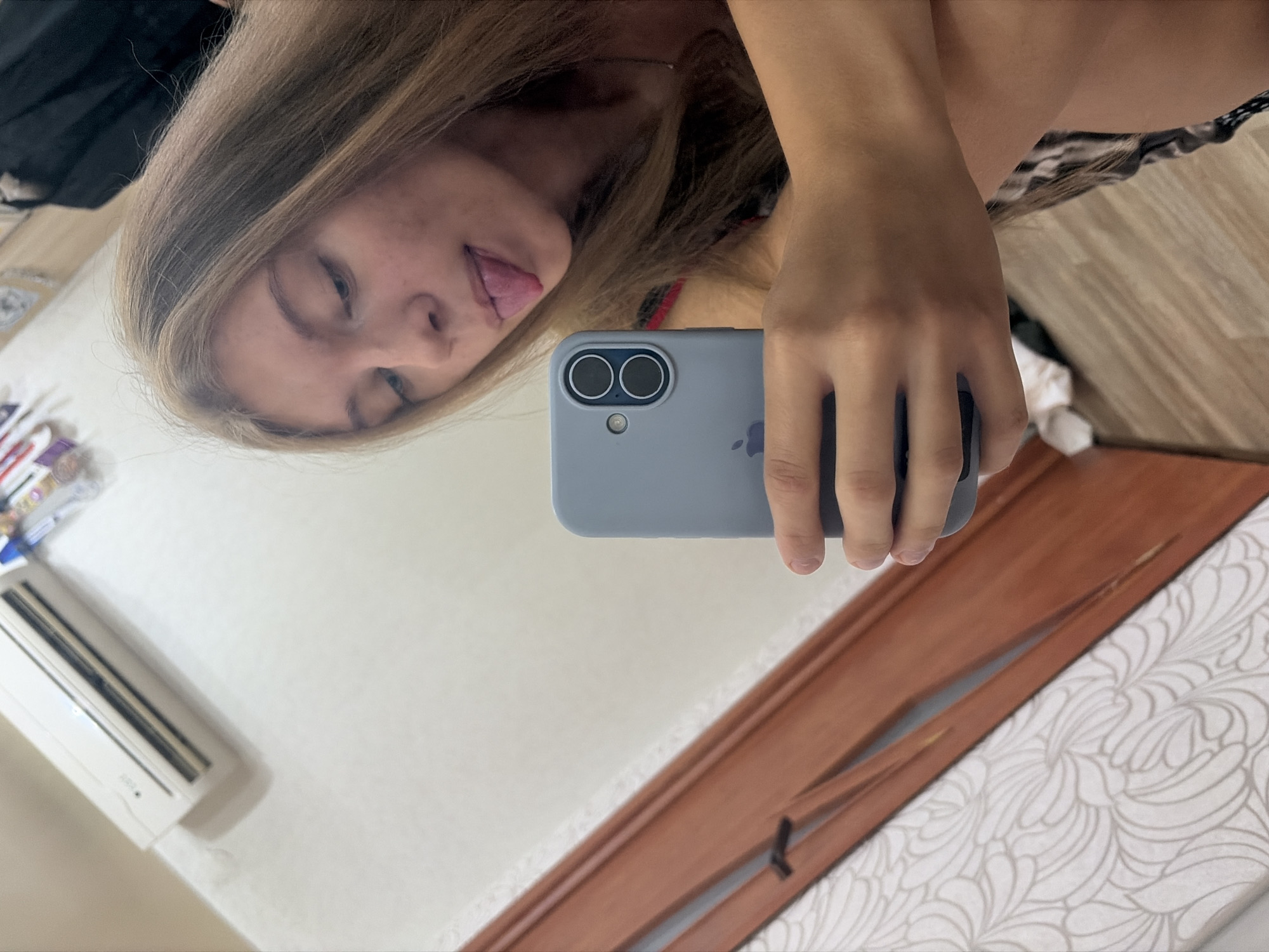 The image captures a selfie taken by a young woman in what appears to be a bedroom. She has long blonde hair and is sticking her tongue out at the camera. She is holding a light blue smartphone in her right hand. In the background, an air conditioner is visible on the wall, and to the left, a wooden frame is present. The flooring appears to be wood. The overall impression is an informal snapshot within a residential setting. There is no direct indication of the city of Chaikovsky, Russia, within the image itself.