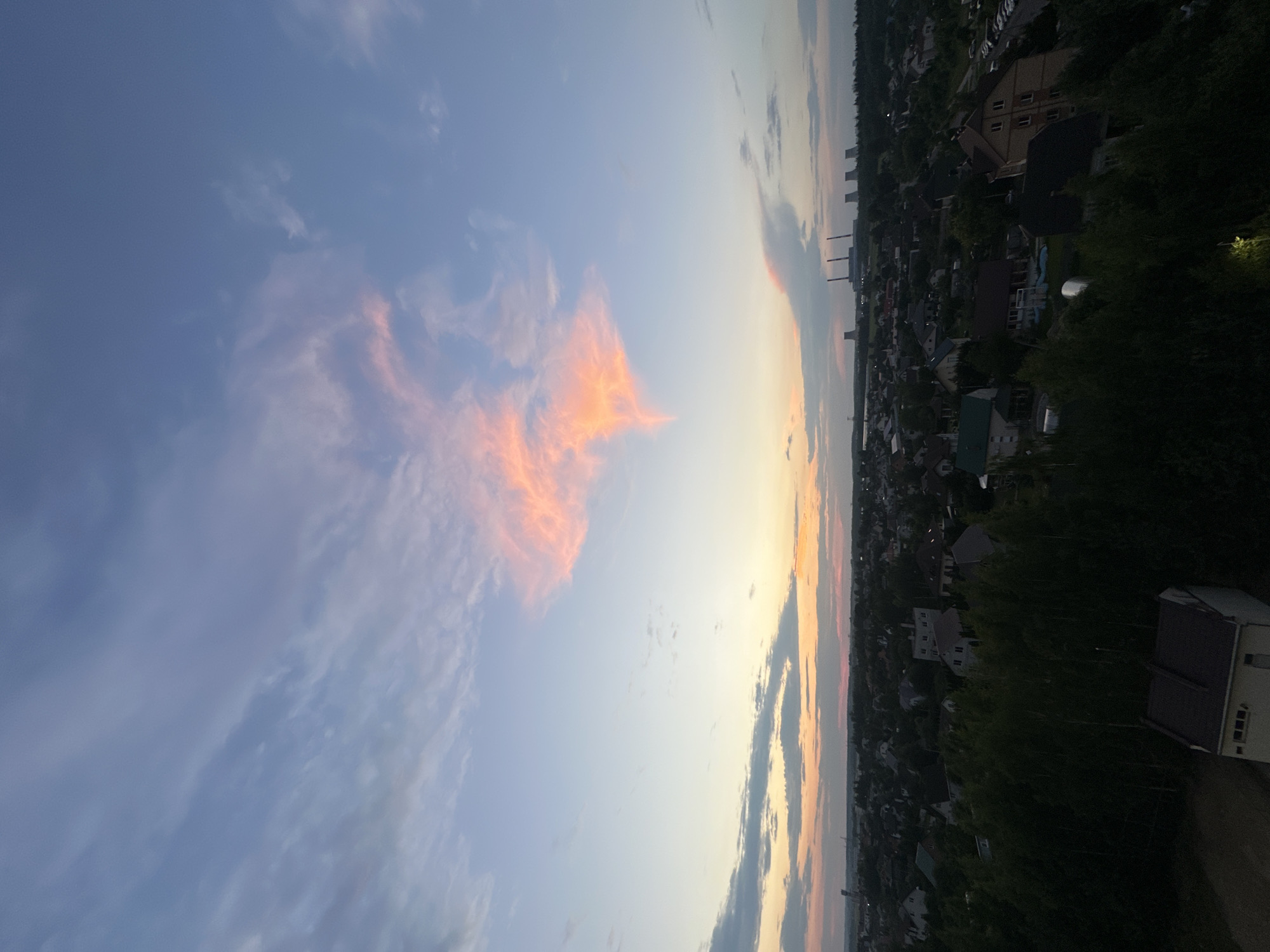 This is an aerial photograph taken from a high vantage point overlooking a suburban area that appears to be located in the vicinity of the Shchomyslitsky Sel' soviety, Belarus. The image captures a twilight scene with a dramatic sky dominated by vibrant hues of pink, orange, and blue clouds. In the foreground, the dark silhouettes of trees and rooftops of houses are visible, indicating a residential area with dense vegetation. Beyond the immediate neighborhood, a town or city stretches into the distance, characterized by a collection of buildings and what appear to be industrial cooling towers or chimneys in the far background. The overall impression is one of a peaceful, rural or semi-rural landscape under a visually striking sky. There are no people or discernible actions captured in the photograph.