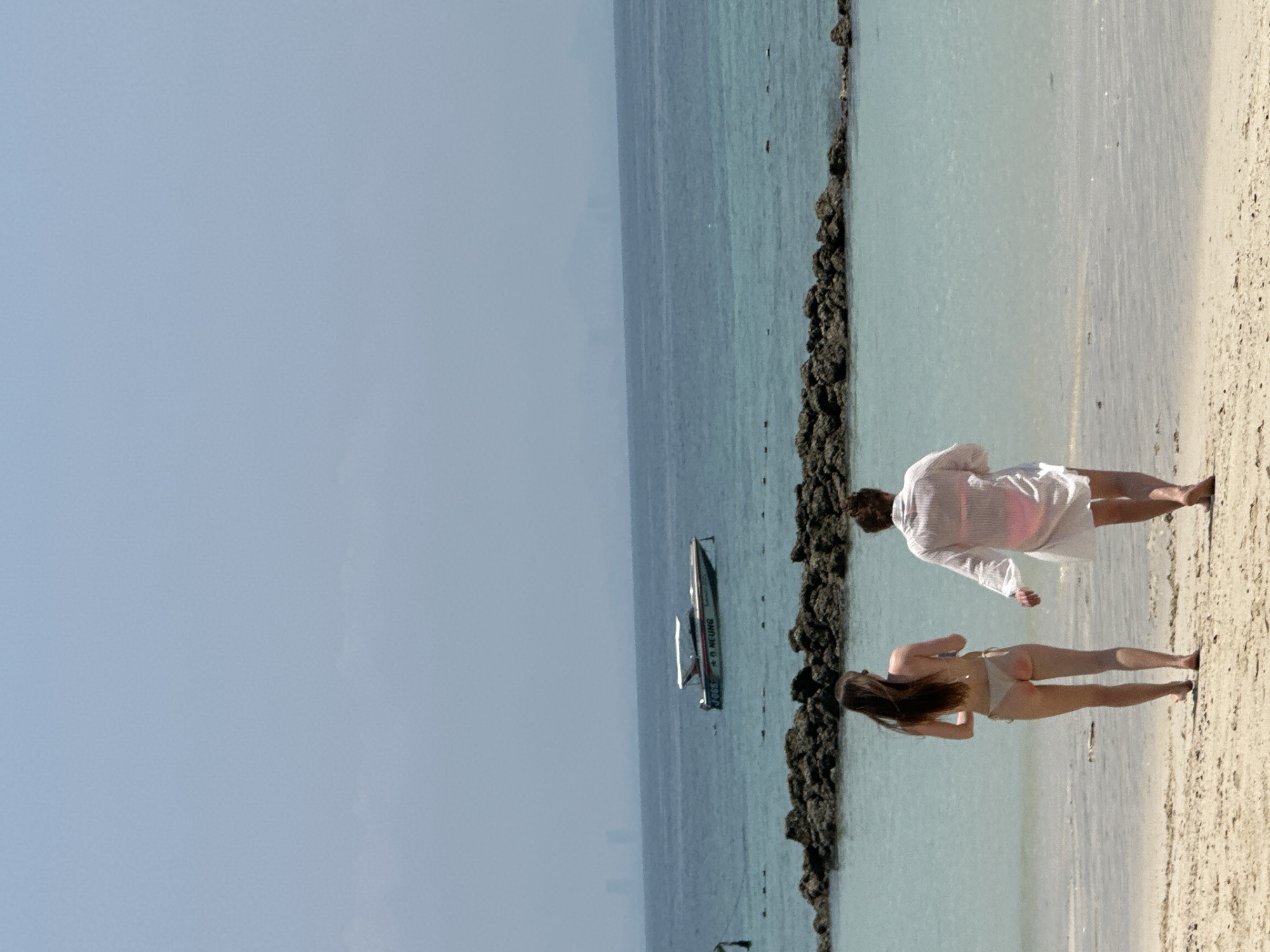 Two people stand on a beach next to the ocean. One person wears a white shirt. The other wears a bathing suit. A boat is in the water. The location is near Pattaya City, Thailand.