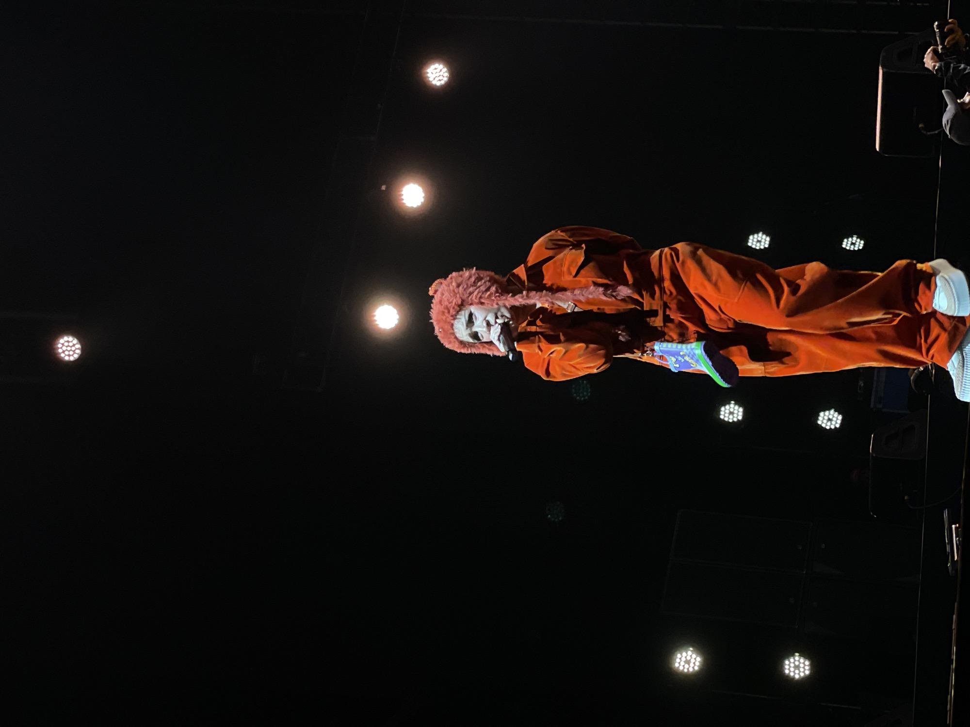 A person is performing on a dark stage with multiple lights. The person is wearing an orange jumpsuit with a pink fuzzy trim around the hood and is holding a microphone to their mouth. They also appear to be wearing white shoes. The setting is a stage at a venue, which may be in Salembaran, Indonesia, although this is only a geographic reference and not integral to the scene. The lights are positioned above and behind the performer, creating a dramatic effect.